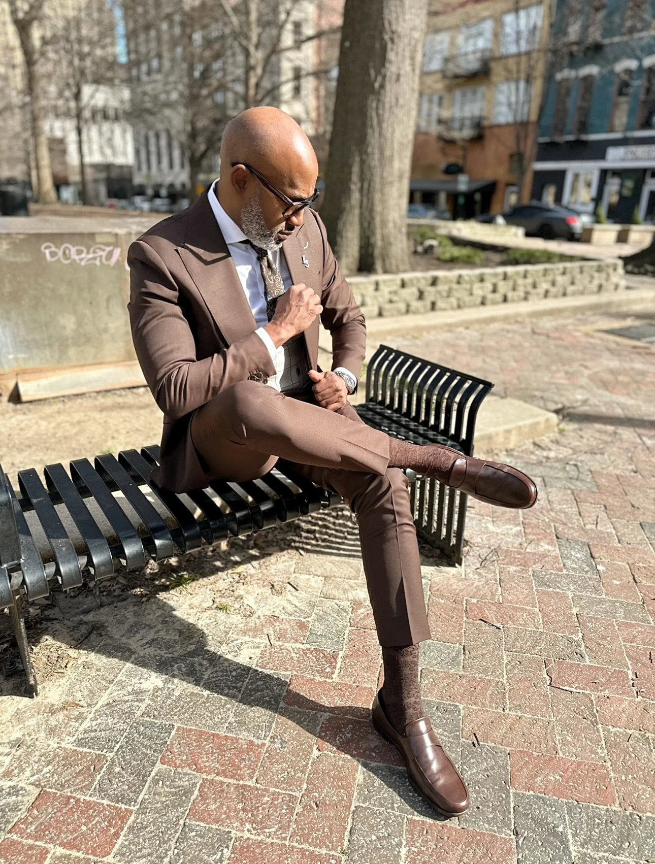 A man in a brown suit, wearing glasses and a watch, sitting on a black park bench outdoors, with one leg crossed over the other, adjusting his sleeve, in a city park with trees and buildings in the background.