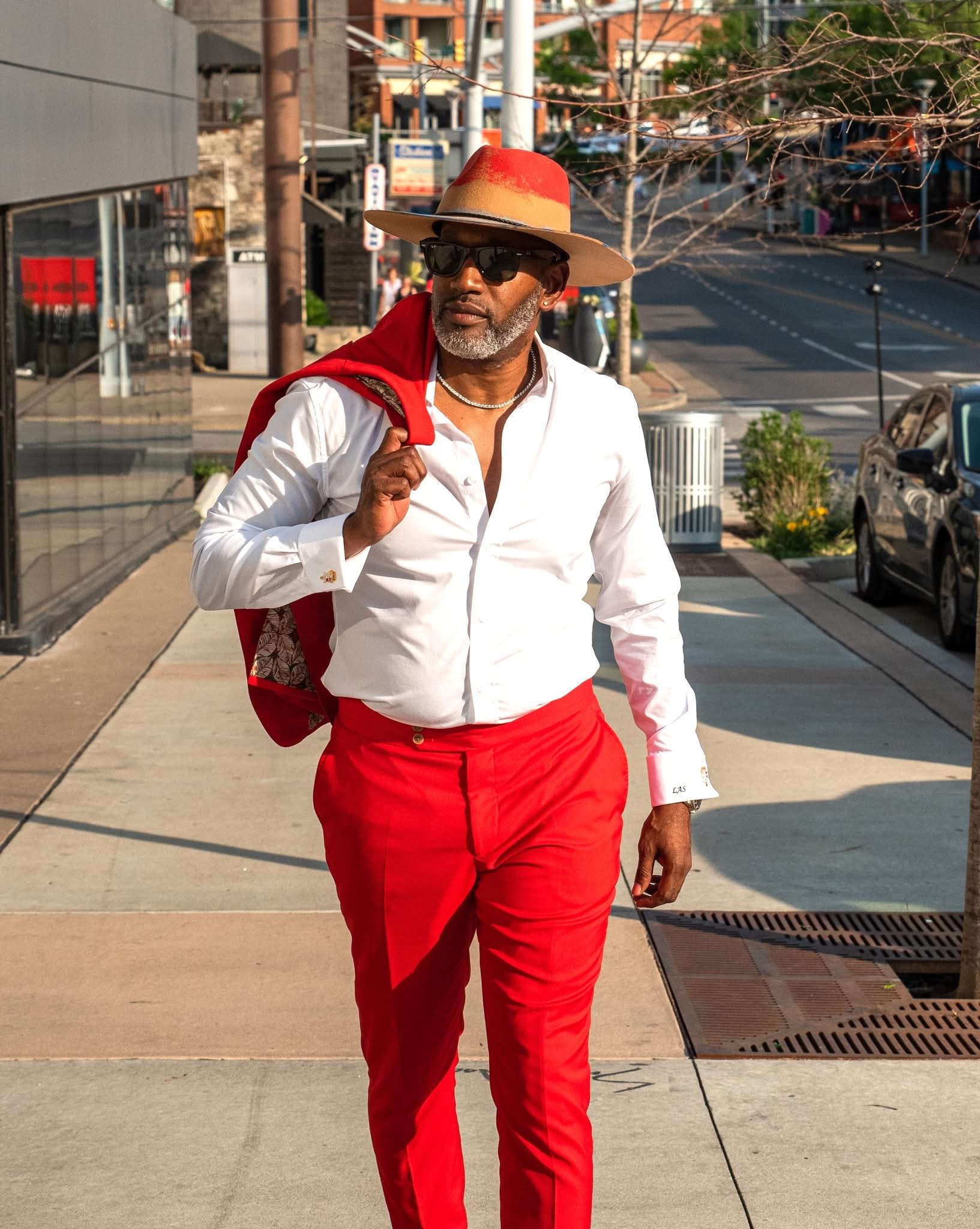 A stylish man walking down the sidewalk in a white shirt, red pants, a red jacket slung over his shoulder, a beige and red fedora, black sunglasses, and a silver necklace.