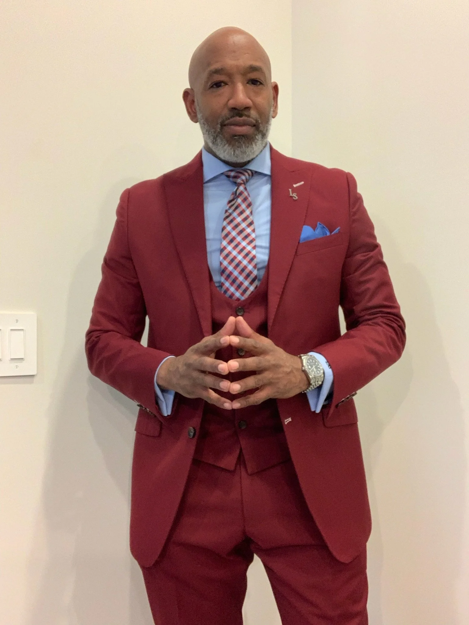 A man in a red suit with a blue shirt, patterned tie, and pocket square. He is standing against a plain beige wall, with hands clasped in front, looking at the camera.