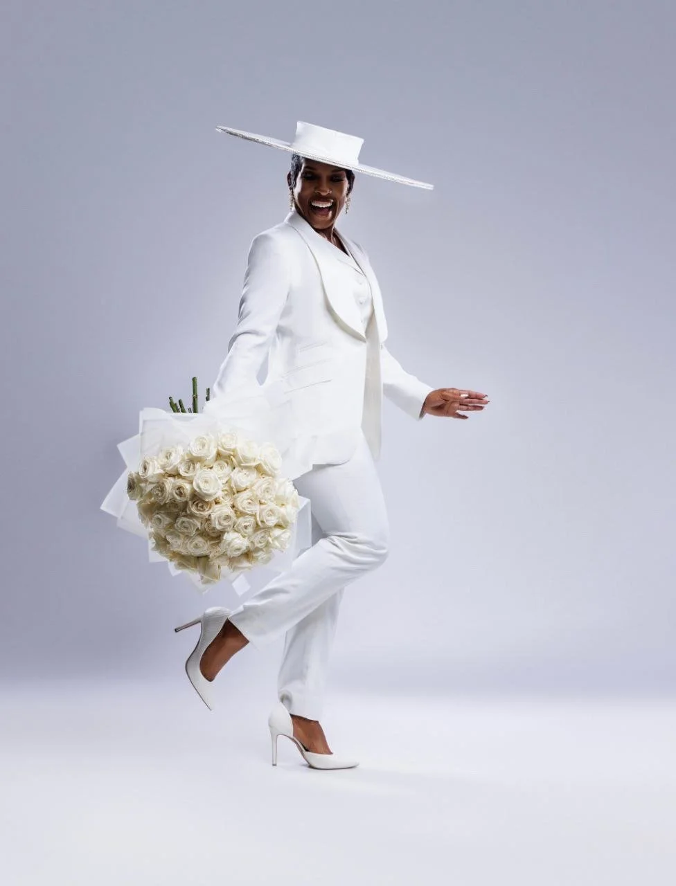 Woman dressed in an all-white outfit, holding a bouquet of white roses and wearing a large white hat, standing on one leg and smiling.