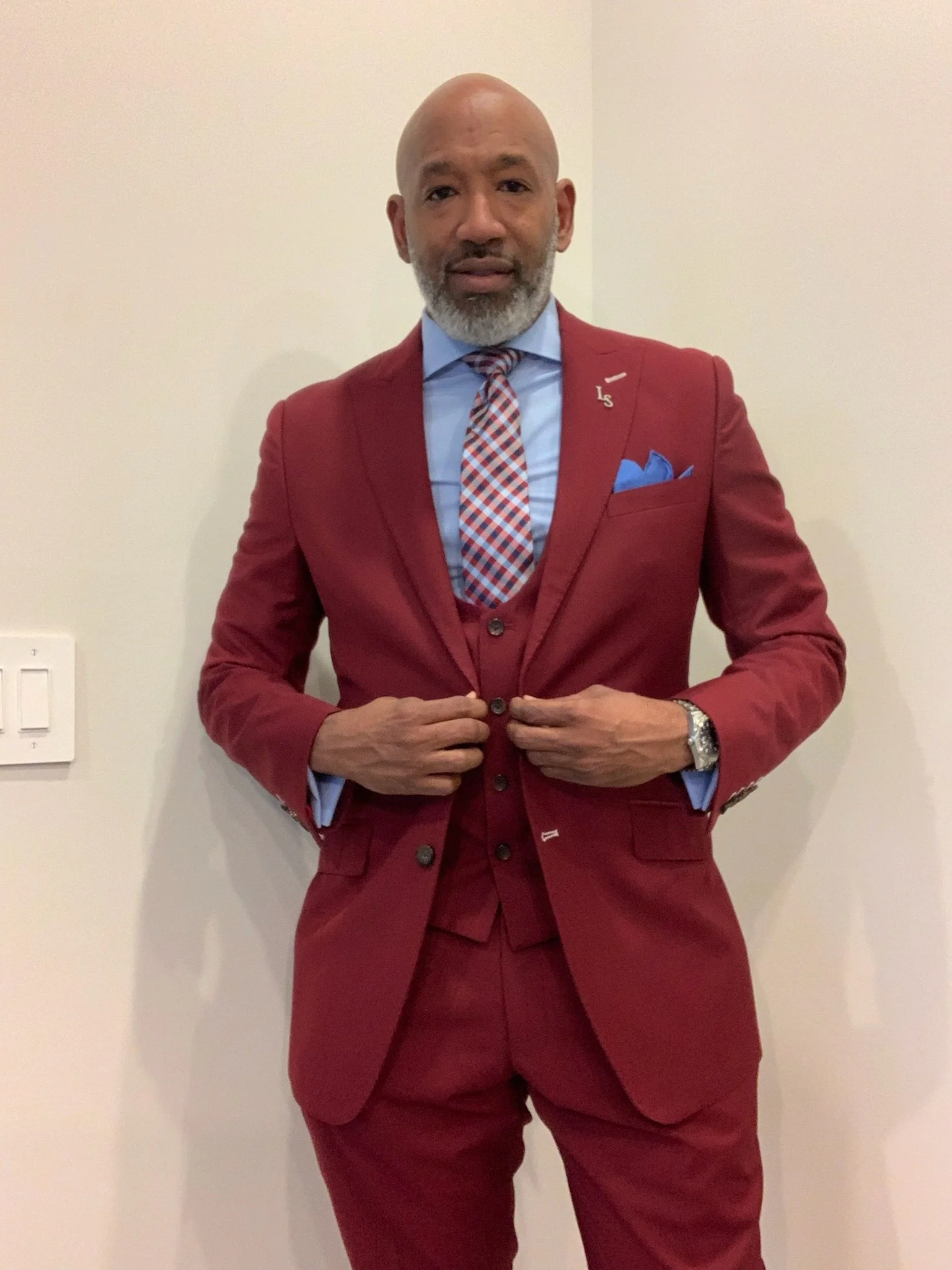 A man wearing a red suit, blue shirt, and a checkered tie, standing in front of a plain white wall. He is adjusting his suit jacket with his hands.