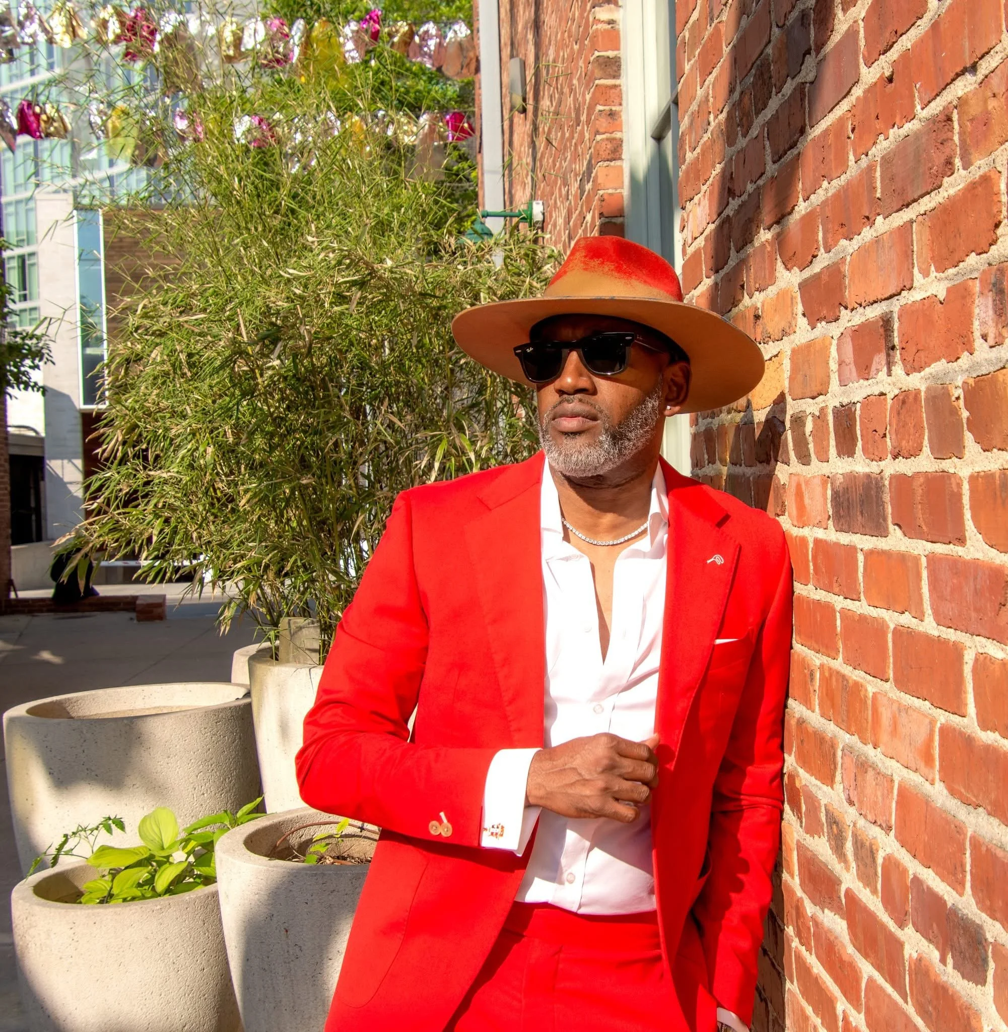 A stylish man in a red suit, white shirt, and black sunglasses, leaning against a brick wall outdoors, wearing a wide-brimmed beige hat with a red band, with potted plants behind him.