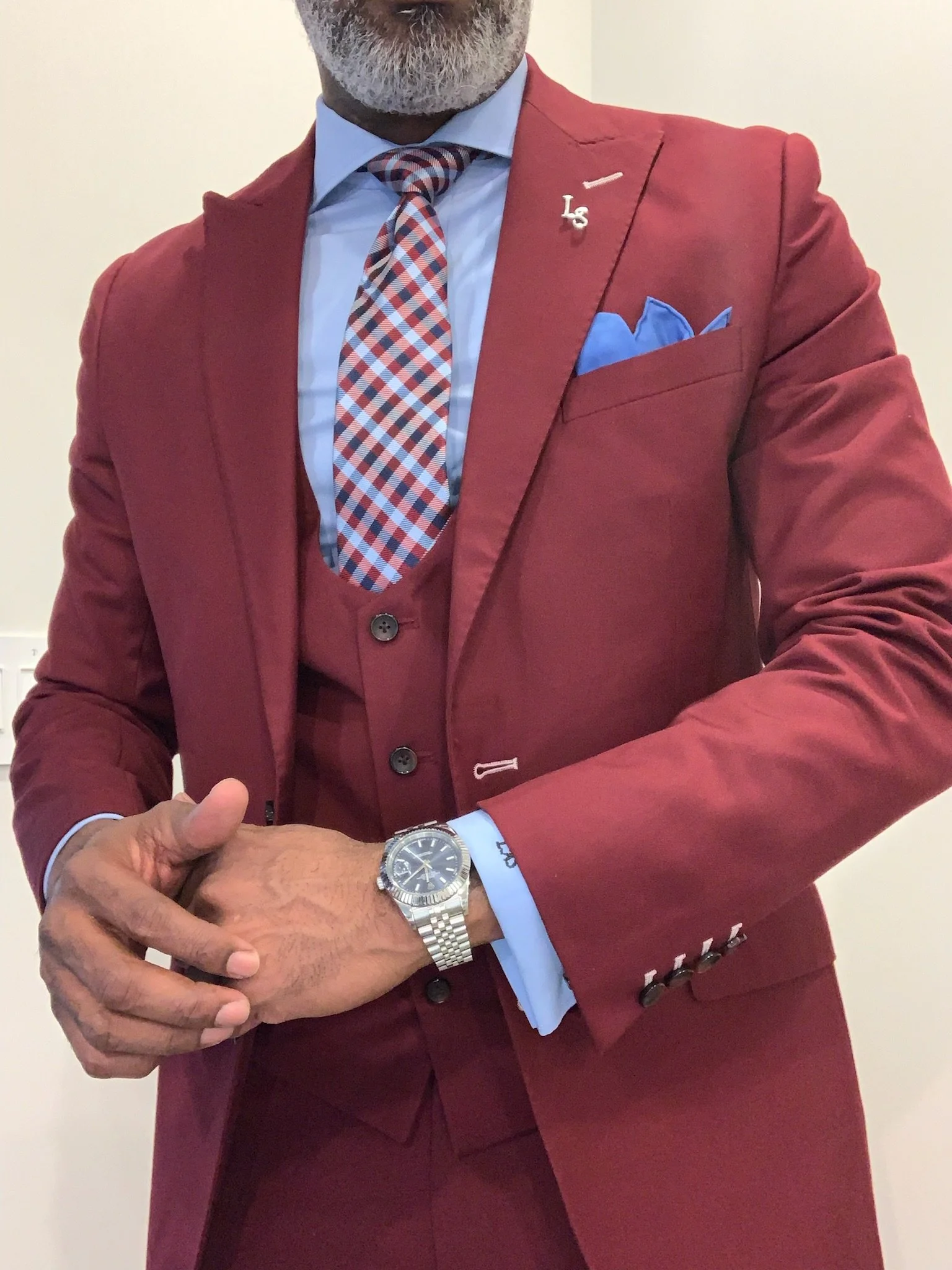 A man dressed in a red suit with a light blue shirt, patterned red, blue, and white tie, and a matching pocket square. He is wearing a silver wristwatch on his left wrist and has a small lapel pin with the initials 'L S' on his jacket.