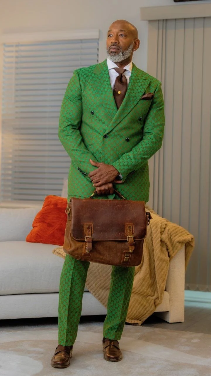 A well-dressed man with a beard stands in a living room holding a brown leather briefcase. He is wearing a green patterned suit with a double-breasted jacket, brown shirt, and a dark tie. There are orange and beige pillows on a white sofa and a beige