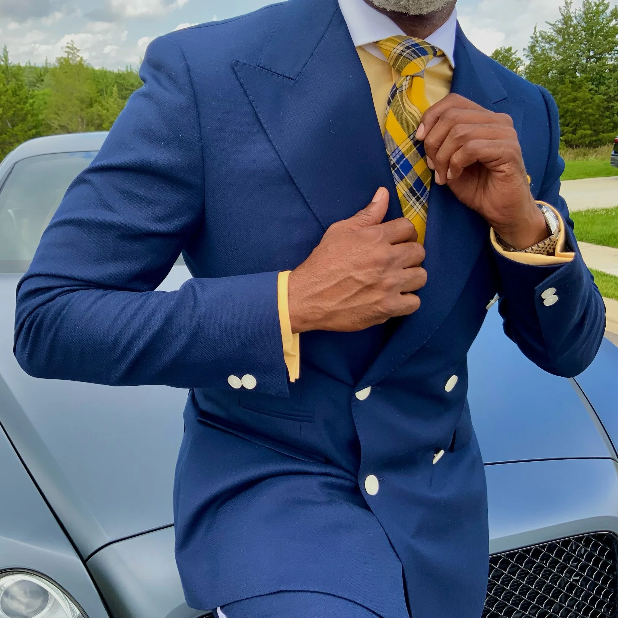 A man in a blue suit adjusting his yellow plaid tie with a silver watch on his left wrist, standing outdoors in front of a parked car and green trees.