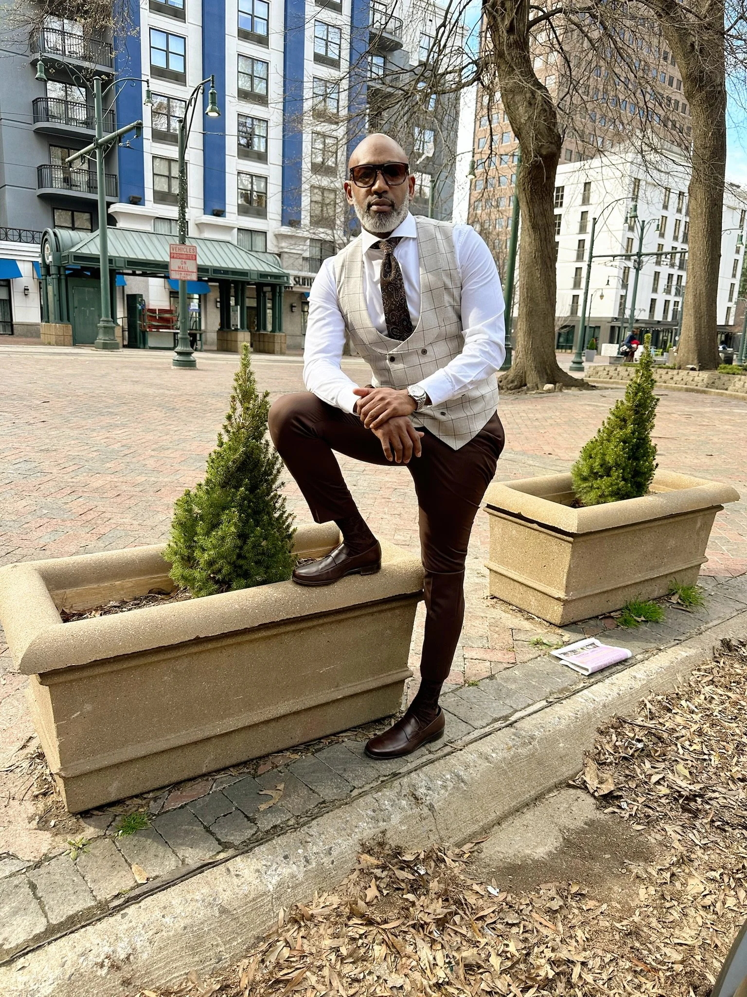A man dressed in a white shirt, beige plaid vest, and brown pants poses with one foot on a large outdoor concrete planter in an urban park. He wears sunglasses, a patterned tie, and a watch, with a cityscape of tall buildings and leafless trees in th