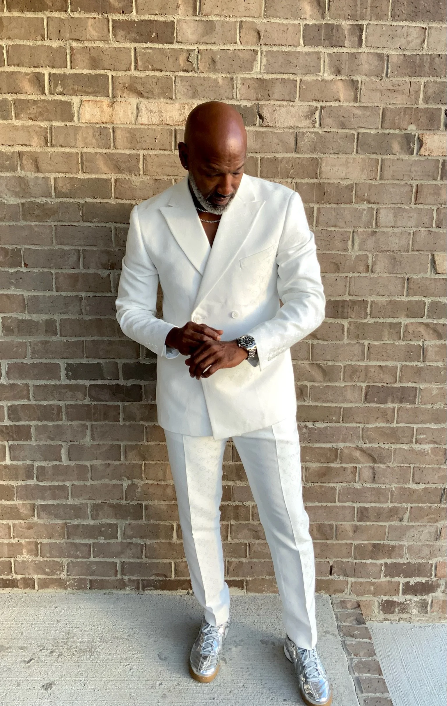 A man dressed in a white suit with a floral pattern, standing against a brick wall, looking down with his hands clasped.