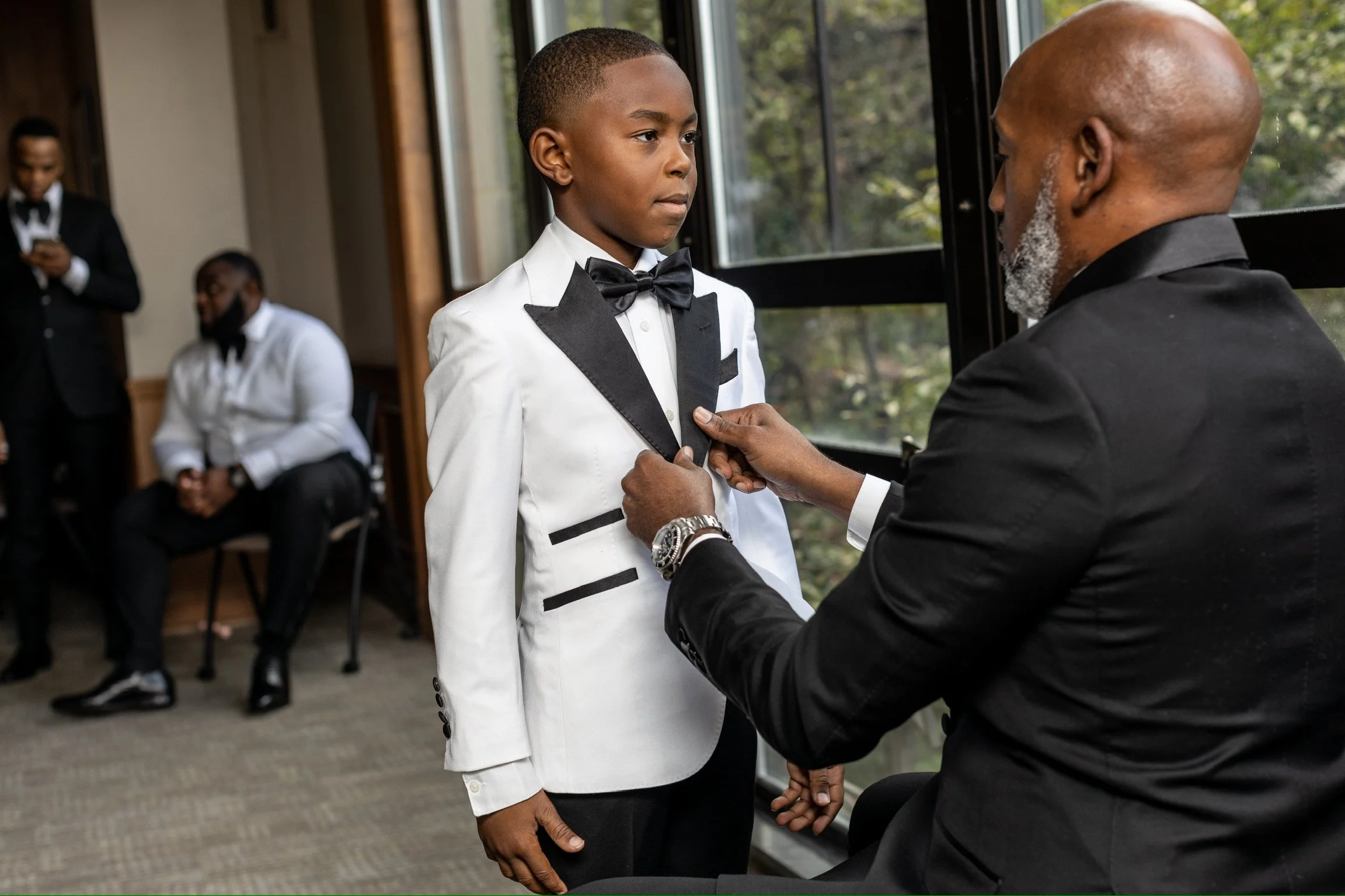 A young boy in a white tuxedo with black lapels and a black bow tie is being helped by an older man in a black tuxedo as he adjusts his tuxedo jacket. Two other men in tuxedos are sitting and standing in the background near a large window, with green