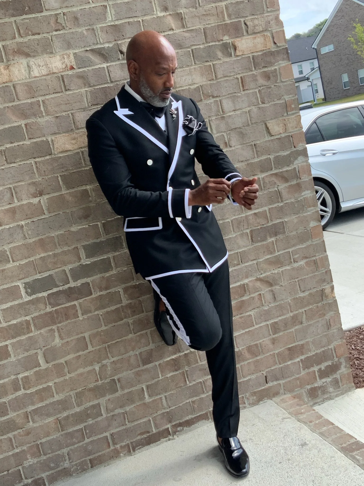 A man dressed in a black tuxedo with white trim and a bow tie, standing against a brick wall outdoors, adjusting his watch.