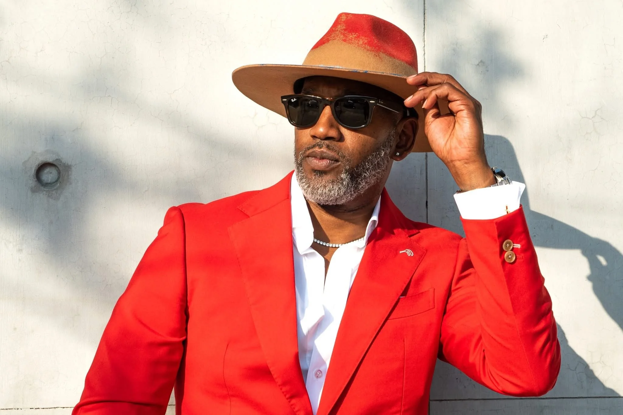 A stylish man wearing a red suit, white shirt, black sunglasses, a beige wide-brimmed hat with red accents, and a silver watch, standing against a white wall with his right hand touching his hat.