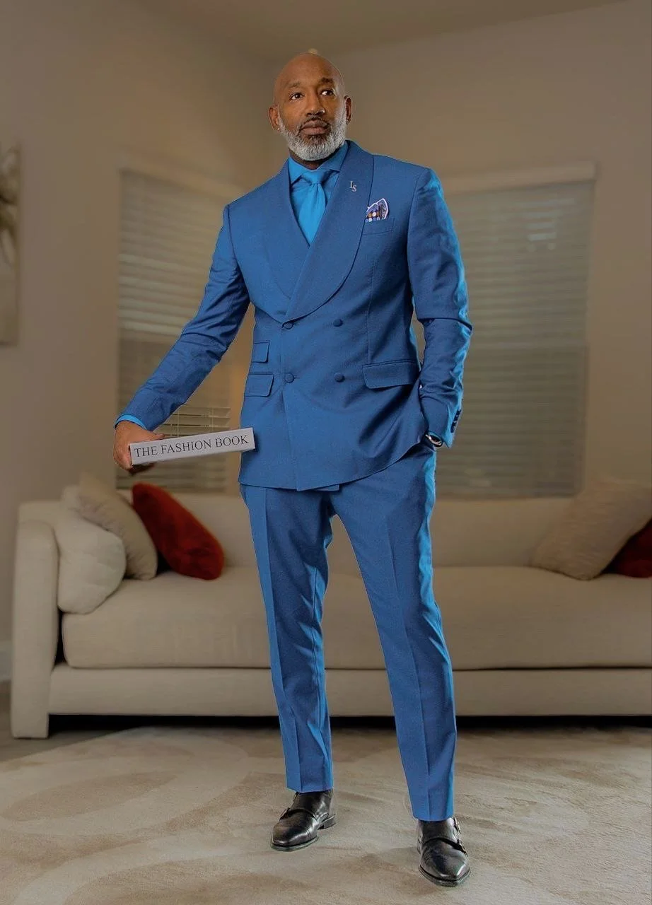A man in a bright blue suit with a matching shirt and tie, standing in a living room holding a book titled "The Fashion Book."