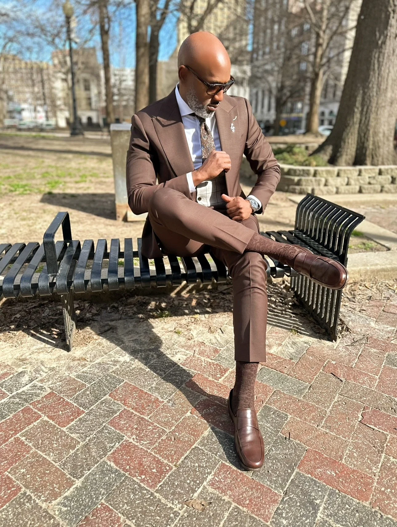A man in a brown suit sitting on a park bench, adjusting his tie, wearing sunglasses, with trees and buildings in the background on a sunny day.
