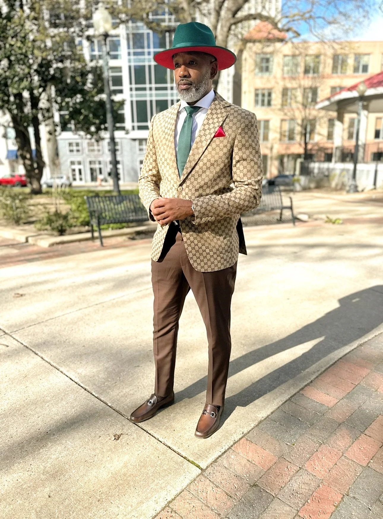 A man in a beige patterned blazer, brown trousers, brown shoes, and a teal and red hat standing outdoors on a sidewalk with benches and trees in the background.