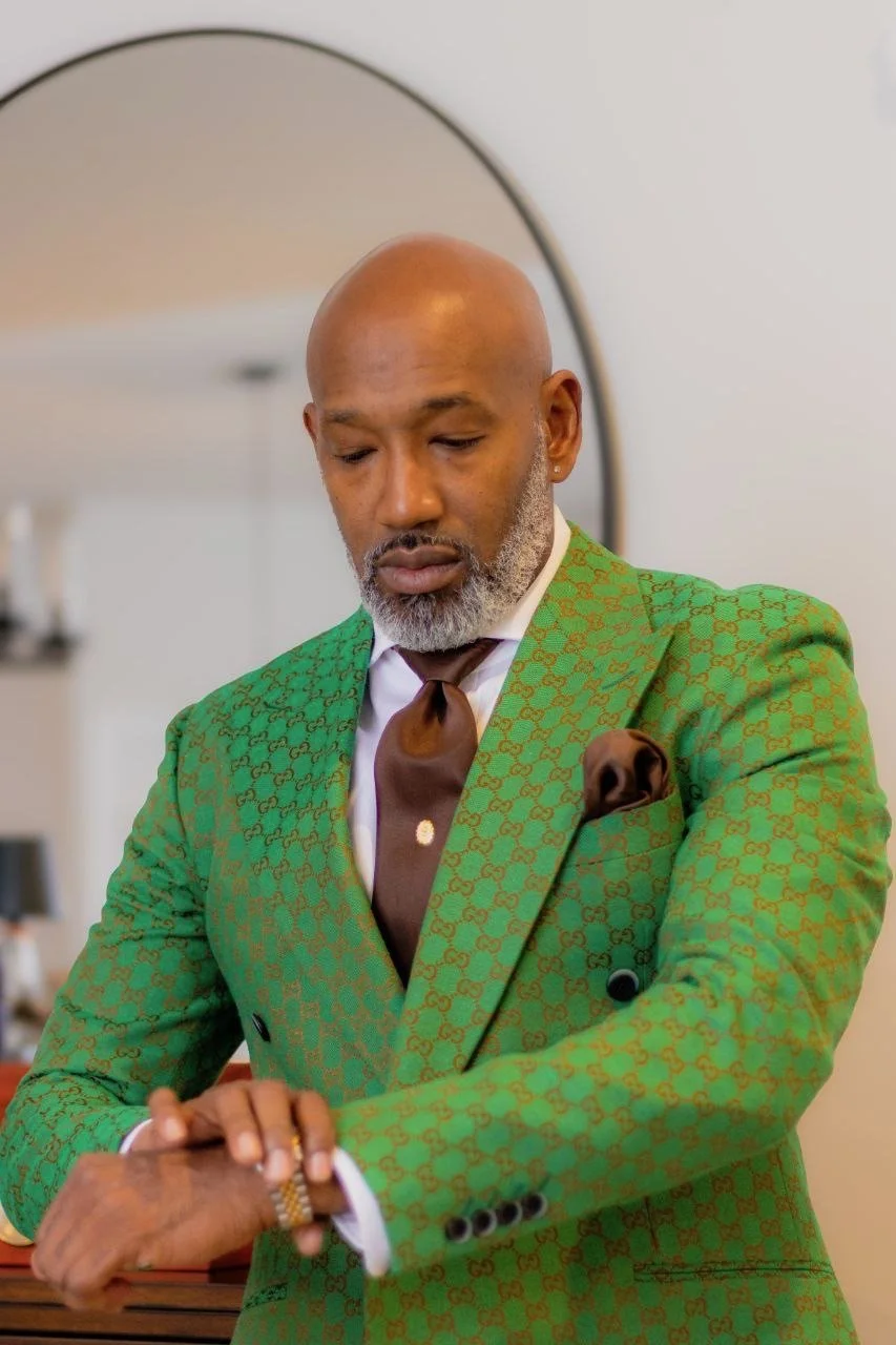 A stylish man with a bald head and gray beard looking at his watch, dressed in a green patterned suit with a brown tie and pocket square, standing indoors in front of a mirror.