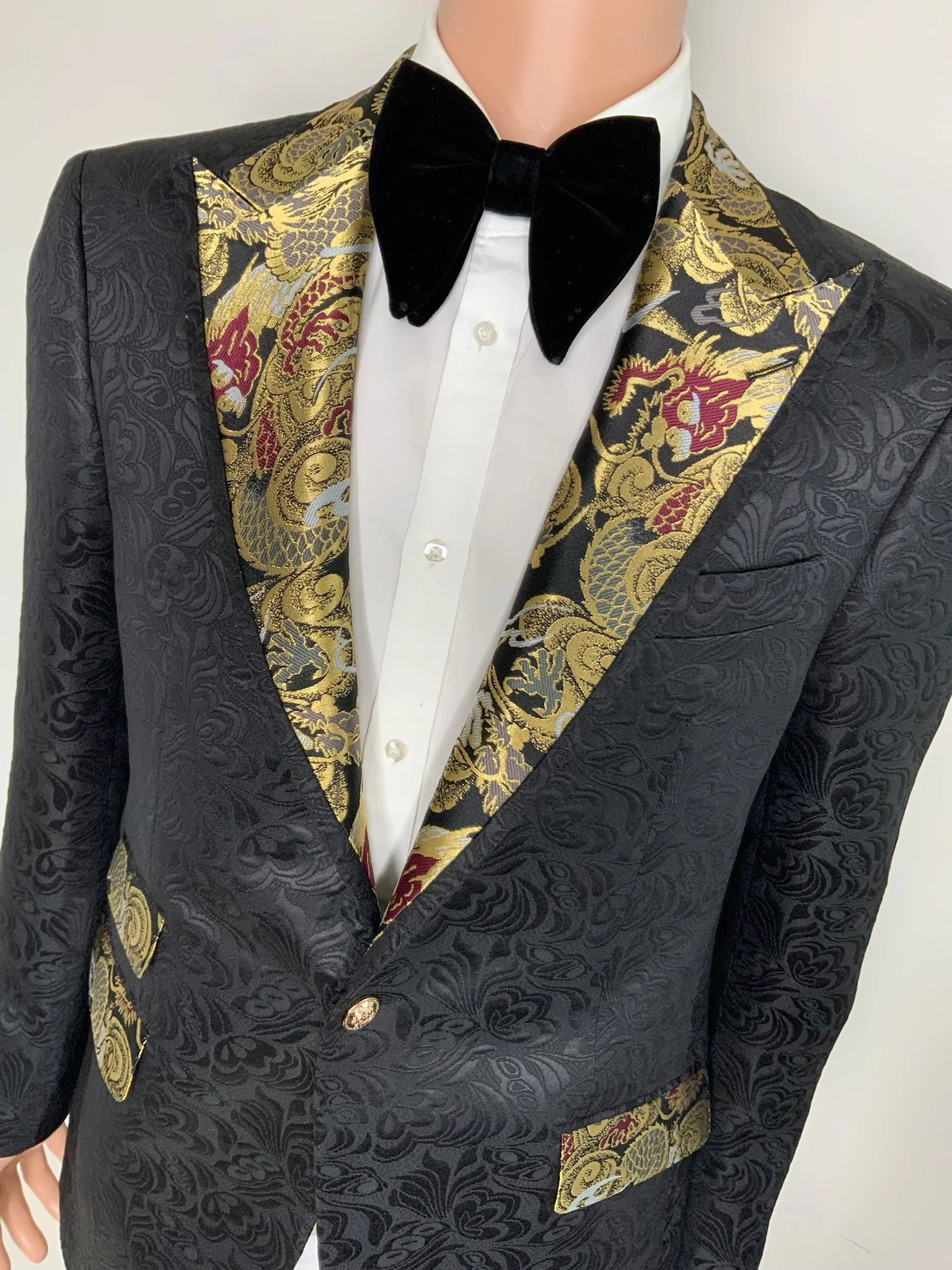 Close-up of a person wearing a black tuxedo with gold ornate details on the lapels, a white dress shirt with pearl buttons, and a black velvet bow tie.