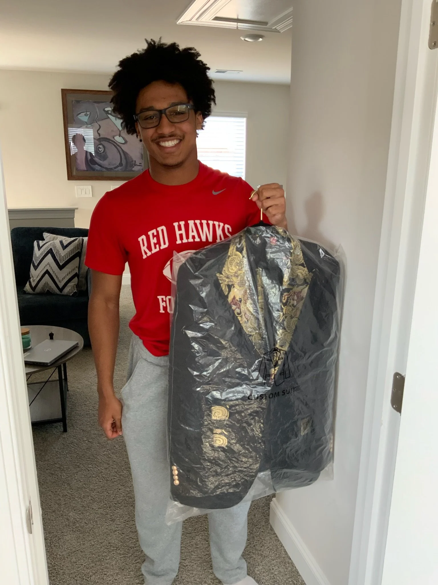A young man with glasses and curly hair smiling, wearing a red 'Red Hawks' shirt and gray sweatpants, holding a suit in a plastic bag.