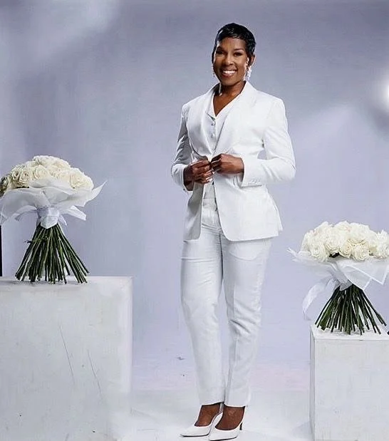 A woman in a white suit standing between two white pedestals with white flower bouquets, smiling at the camera.