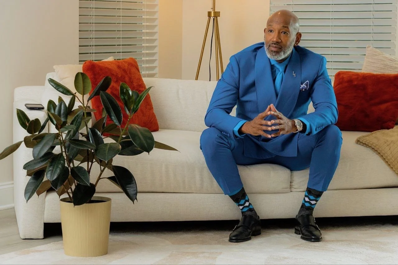A man in a bright blue suit sitting on a cream-colored sofa with orange and beige pillows, in a well-lit room with blinds and a gold floor lamp, next to a large potted plant.