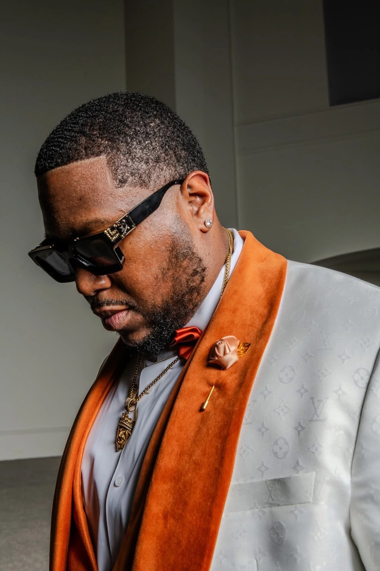 A man wearing stylish black sunglasses, a white shirt, a tan blazer with an orange velvet lapel, a decorative lapel pin, a gold chain, and earrings, looking downward with a neutral expression.
