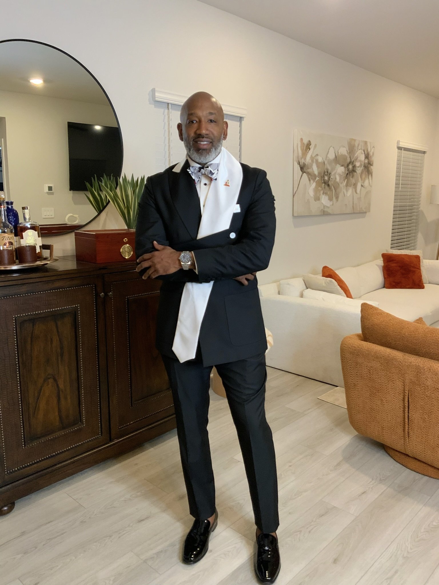 A man in a tuxedo with a white scarf walking in a living room with a large mirror, beige sofa, and abstract wall art.