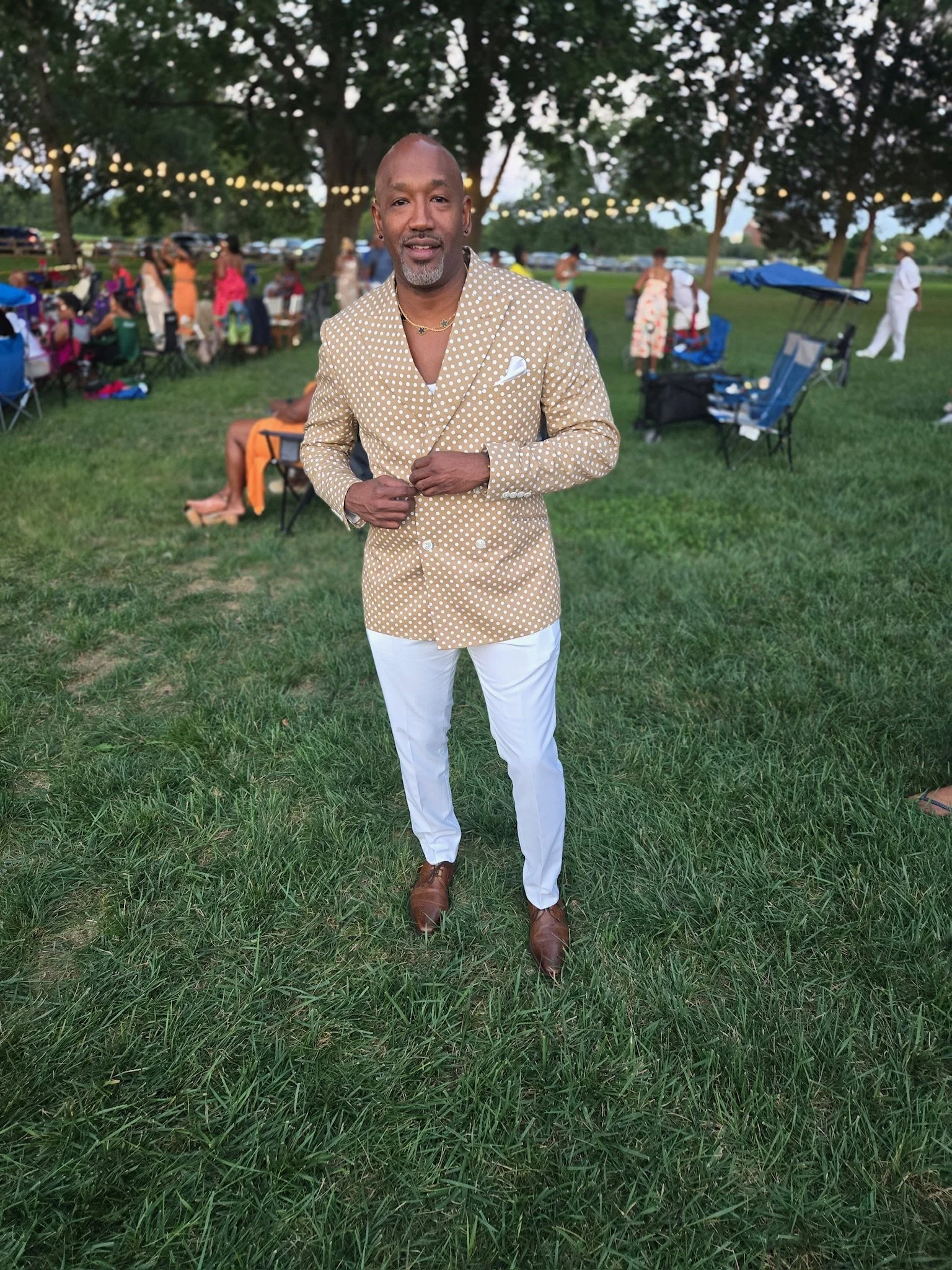 A man in a beige polka dot blazer, white pants, and brown shoes standing on grass at an outdoor event with tables, chairs, and string lights in the background.