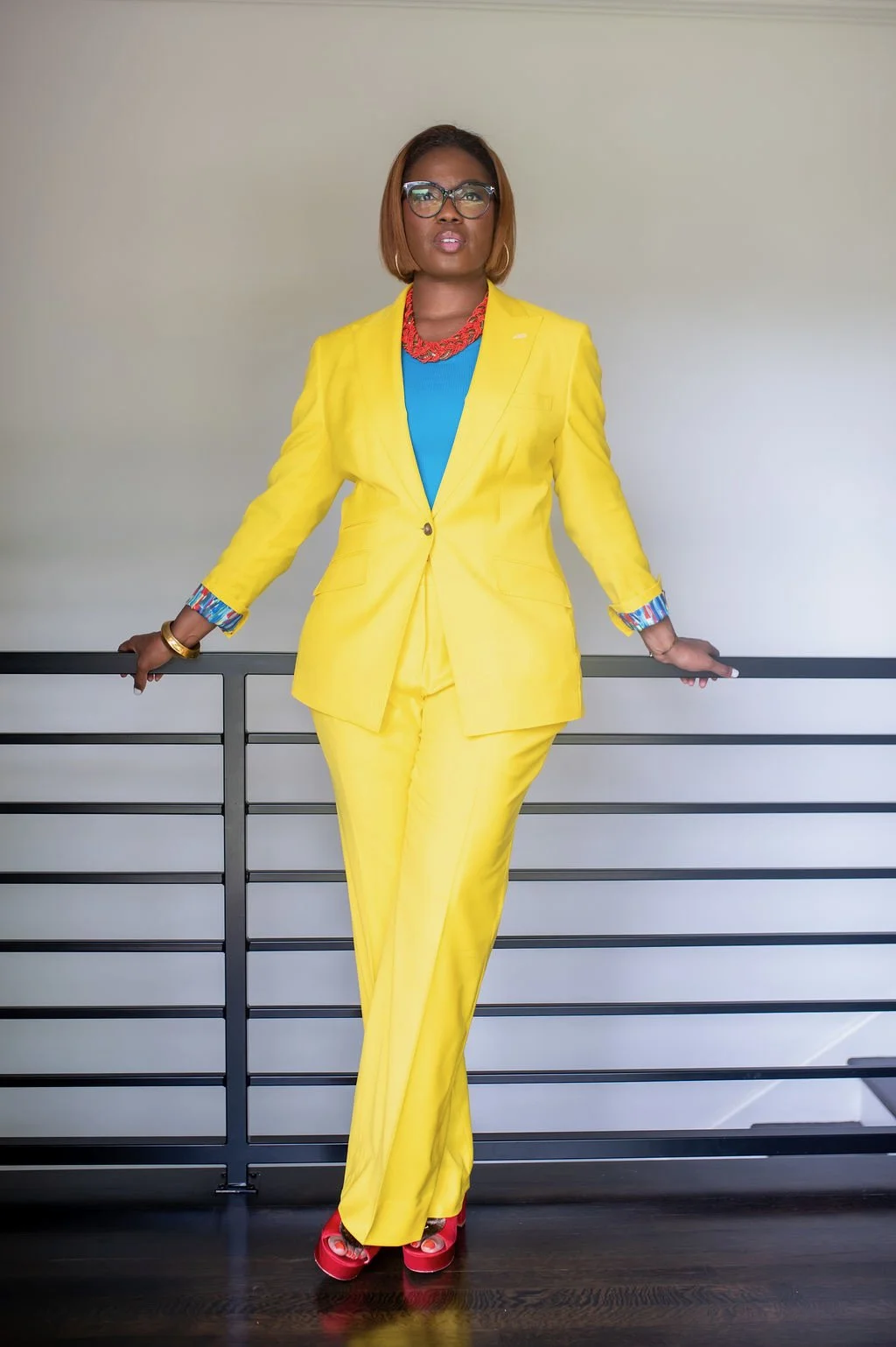 Woman with short hair and glasses wearing a bright yellow suit, blue top, red necklace, and red platform shoes, standing against a neutral background.