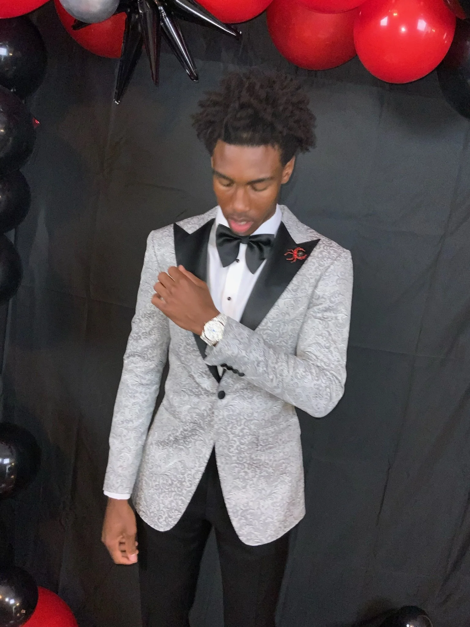 A young man wearing a patterned tuxedo jacket, black bow tie, white dress shirt, and black pants, with a watch on his left wrist, standing against a black background decorated with black, red, and gray balloons.