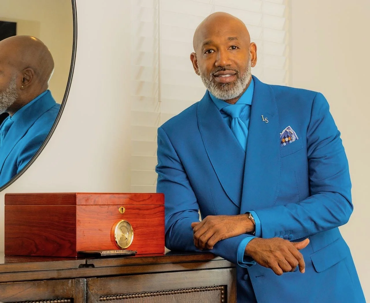 A well-dressed man in a bright blue suit and tie, with a pocket square, standing next to a wooden dresser and a small gold clock, with a mirror reflecting part of his head and background, in a bright room with white blinds.