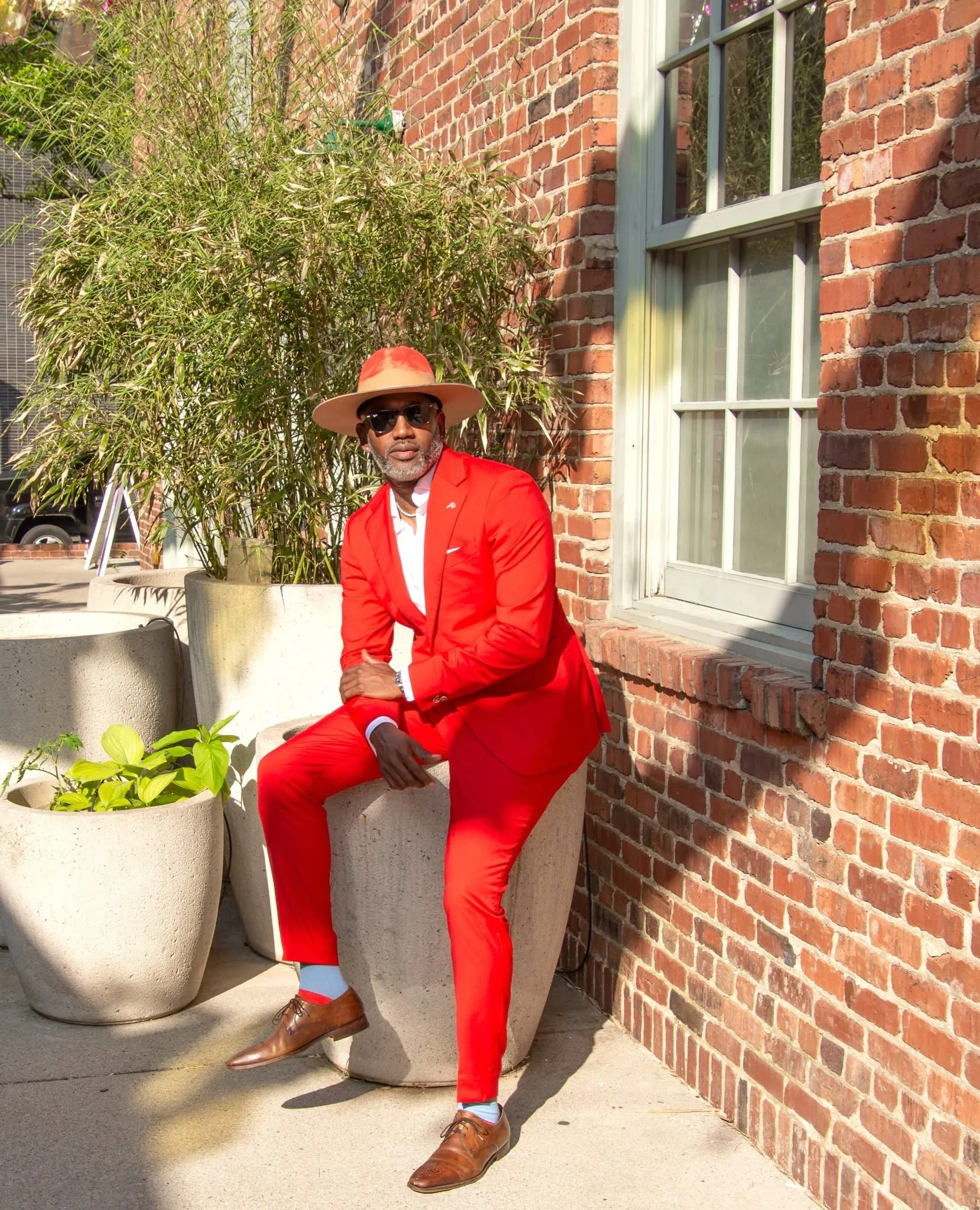 A man dressed in a red suit and hat, wearing sunglasses, sits on a concrete planter outside a brick building with large windows, with greenery in the background.