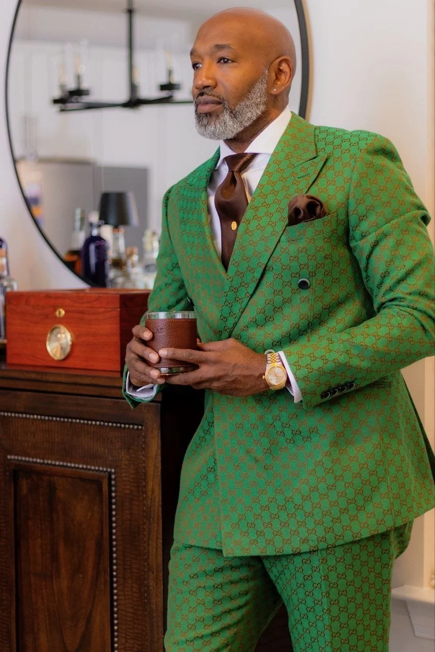 A man with a grey beard dressed in a stylish green suit with a pattern, holding a glass, standing in front of a mirror in a room with a wooden dresser and various bottles.