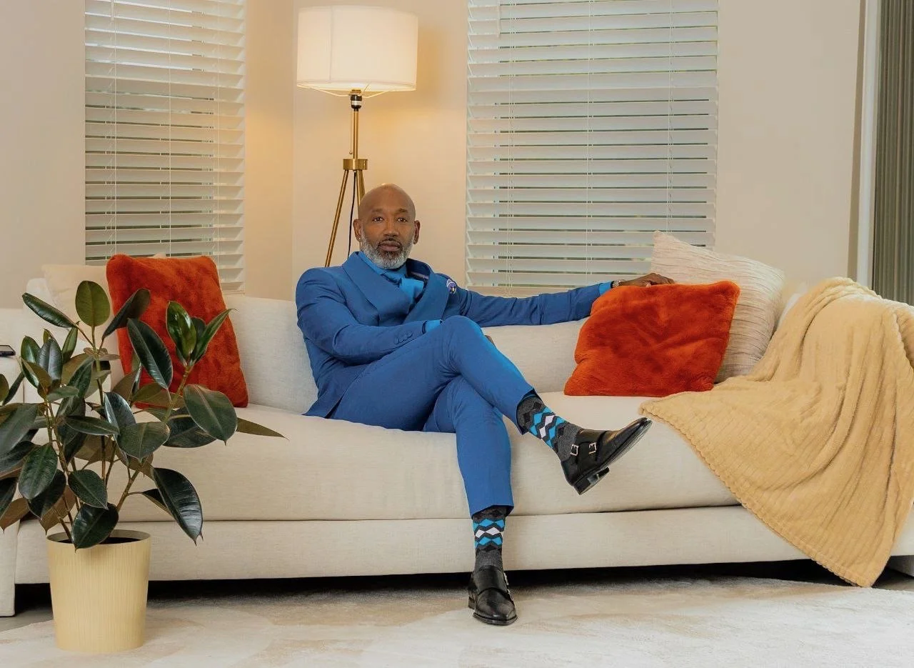 A man in a blue suit sitting on a white couch with orange and cream pillows in a living room with white blinds and a beige blanket. There is a potted plant on the left and a standing lamp behind the man.