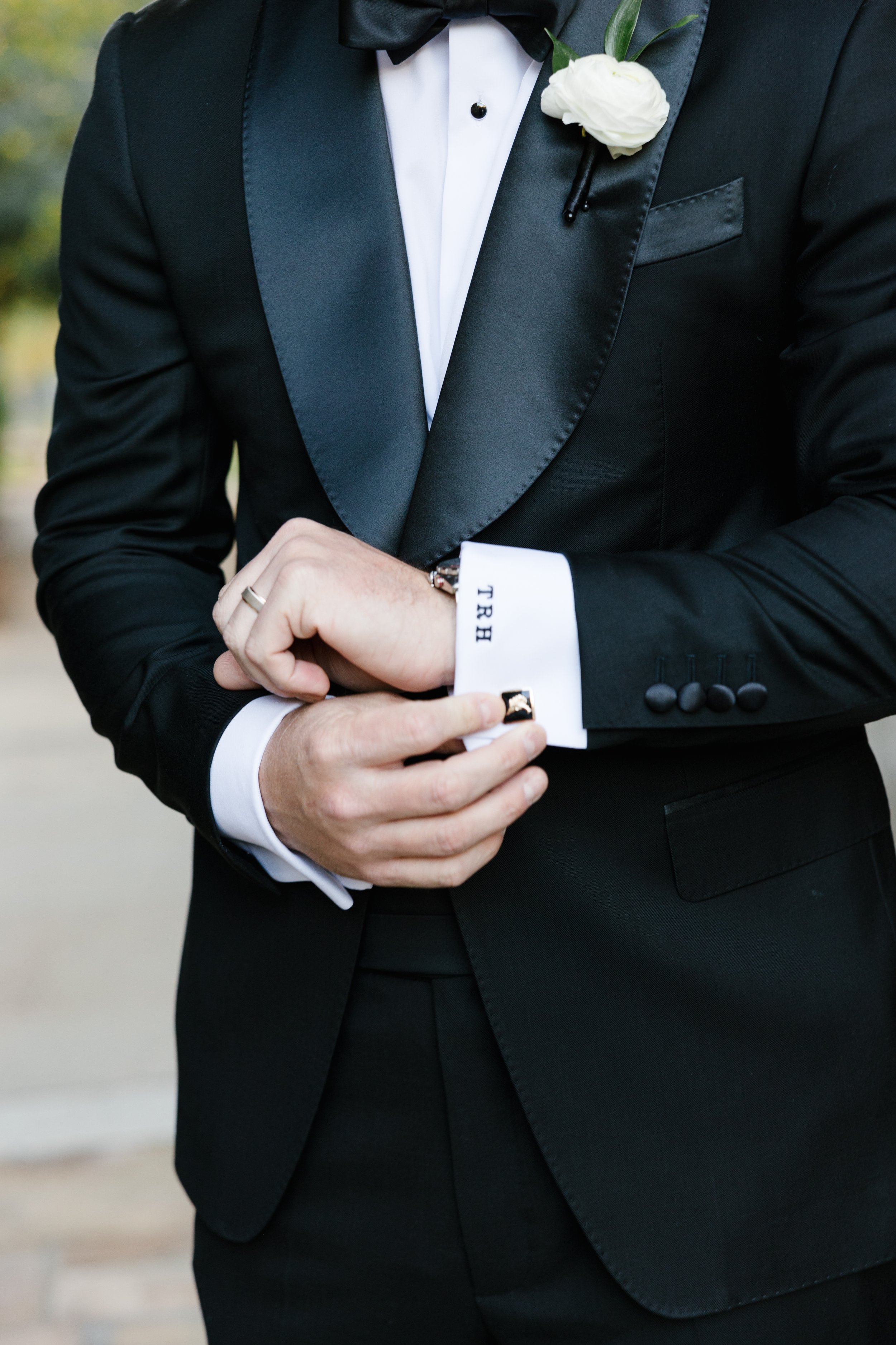 Close-up of a man dressed in a black tuxedo with satin lapels, white dress shirt, and black bow tie, adjusting his cufflink with a white cuff featuring black letters.