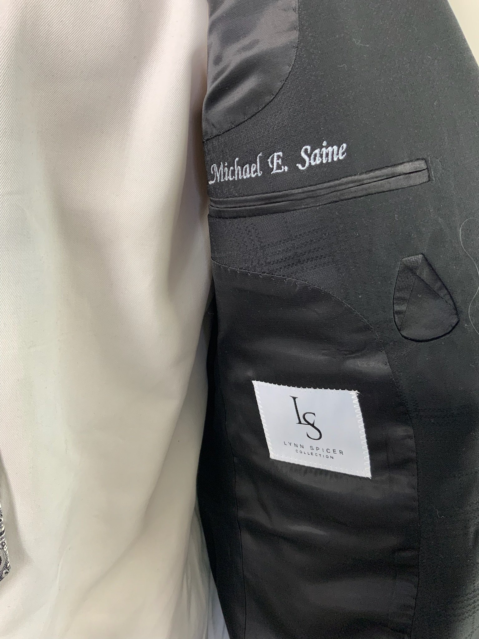 Close-up photo of the inside of a suit jacket showing the lining with embroidered name 'Michael E. Saime' and a branded tag 'Lynn Spicer Collection'.