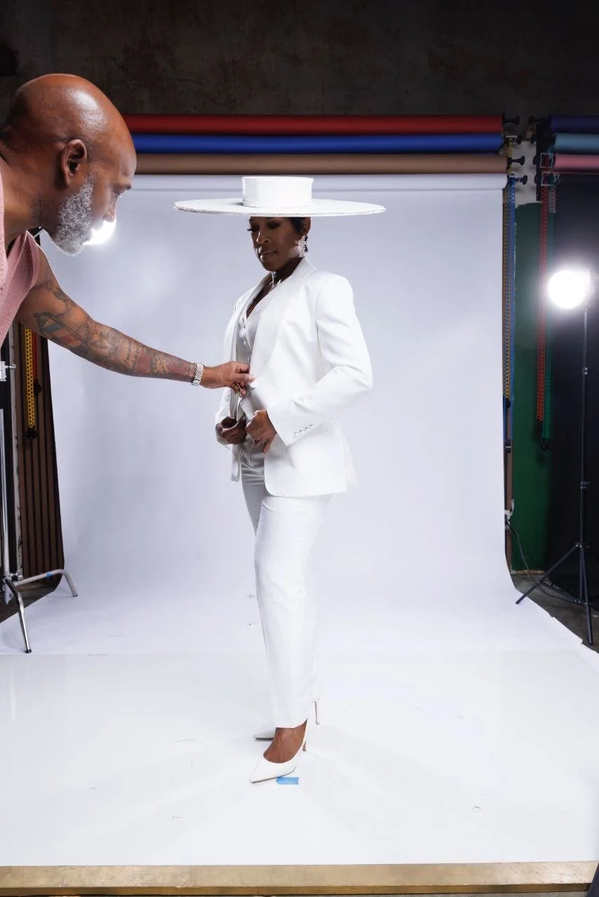 A woman dressed in a white suit with a large white hat standing in a photography studio while a man adjusts her suit.