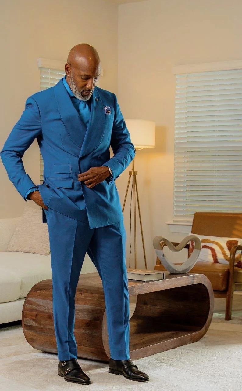 A man in a bright blue suit and black dress shoes adjusting his jacket inside a living room with beige walls, a wooden coffee table, a cream-colored sofa, a wooden chair with a patterned cushion, and a decorative sculpture on the table.