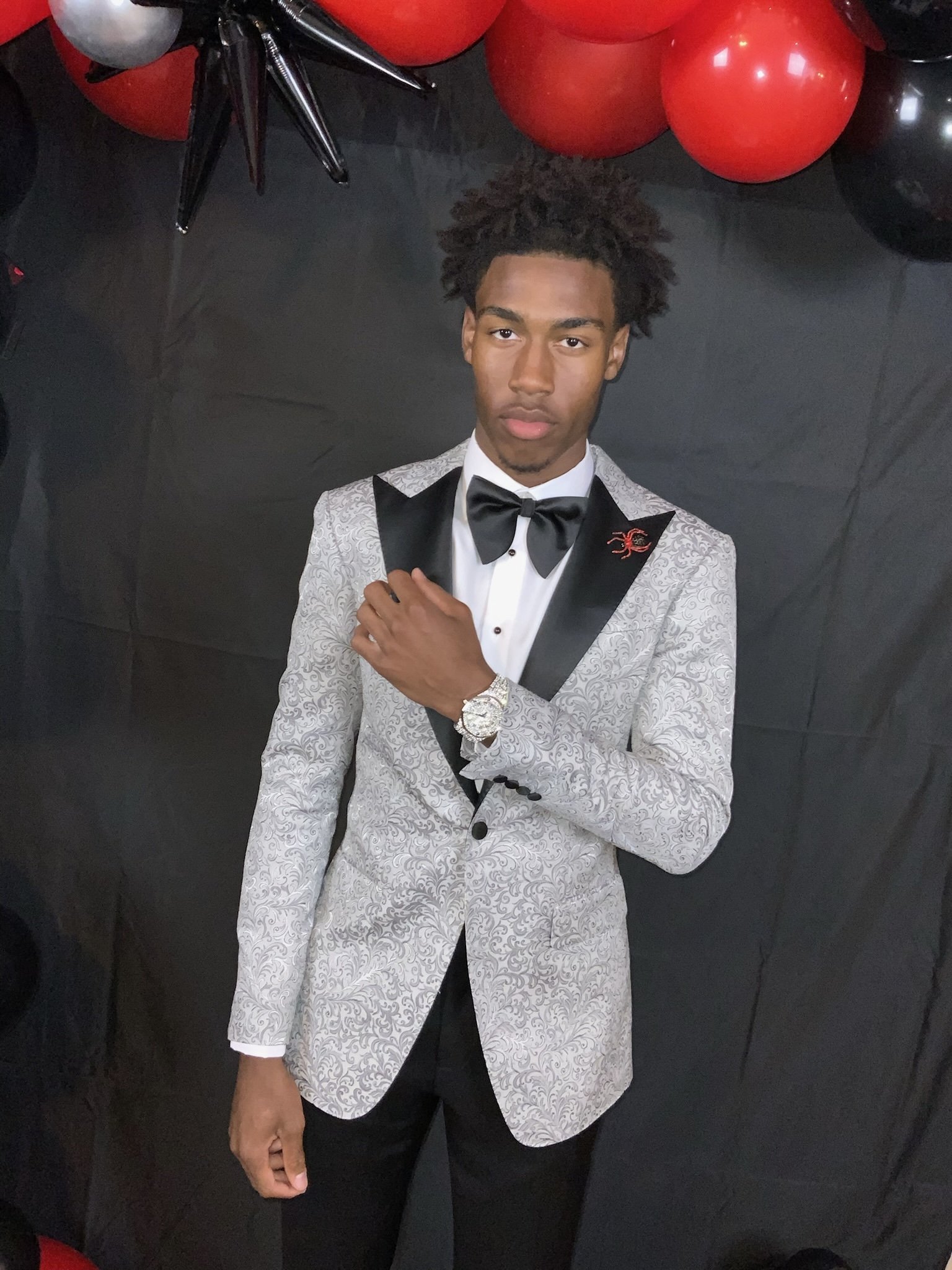 Young man in a white and black tuxedo with a bow tie, standing against a black backdrop with red and black balloons overhead, wearing a watch on his left wrist.