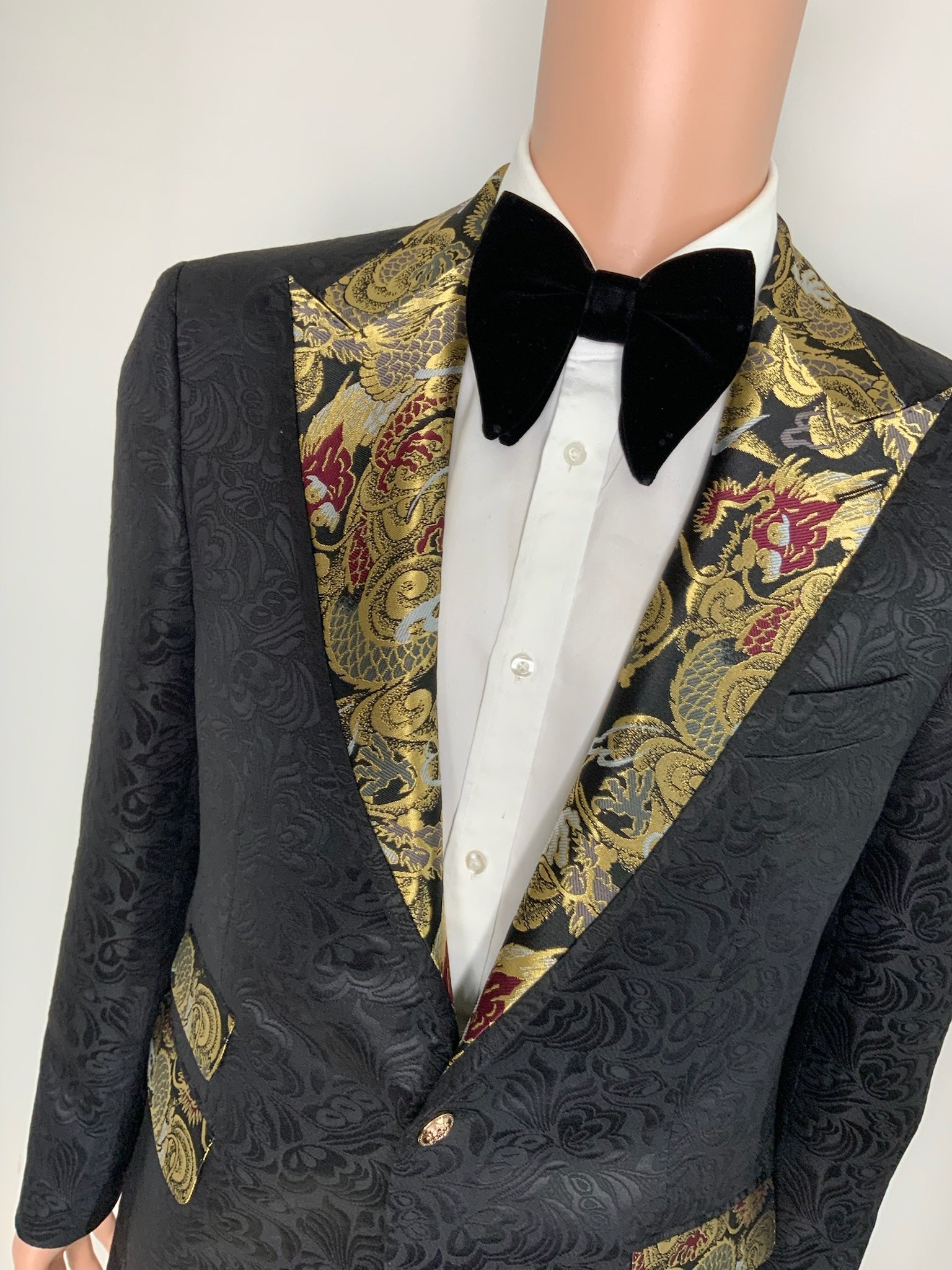 Close-up of a tuxedo with a black floral jacquard pattern, gold and red embroidered lapel accents, a white dress shirt, and a black velvet bow tie on a mannequin.