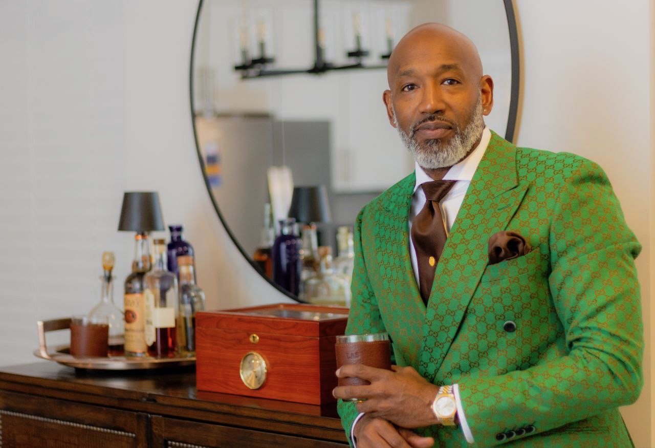 A man with a beard dressed in a green patterned suit holding a glass standing in front of a mirror with bottles and lamps on a wooden table.