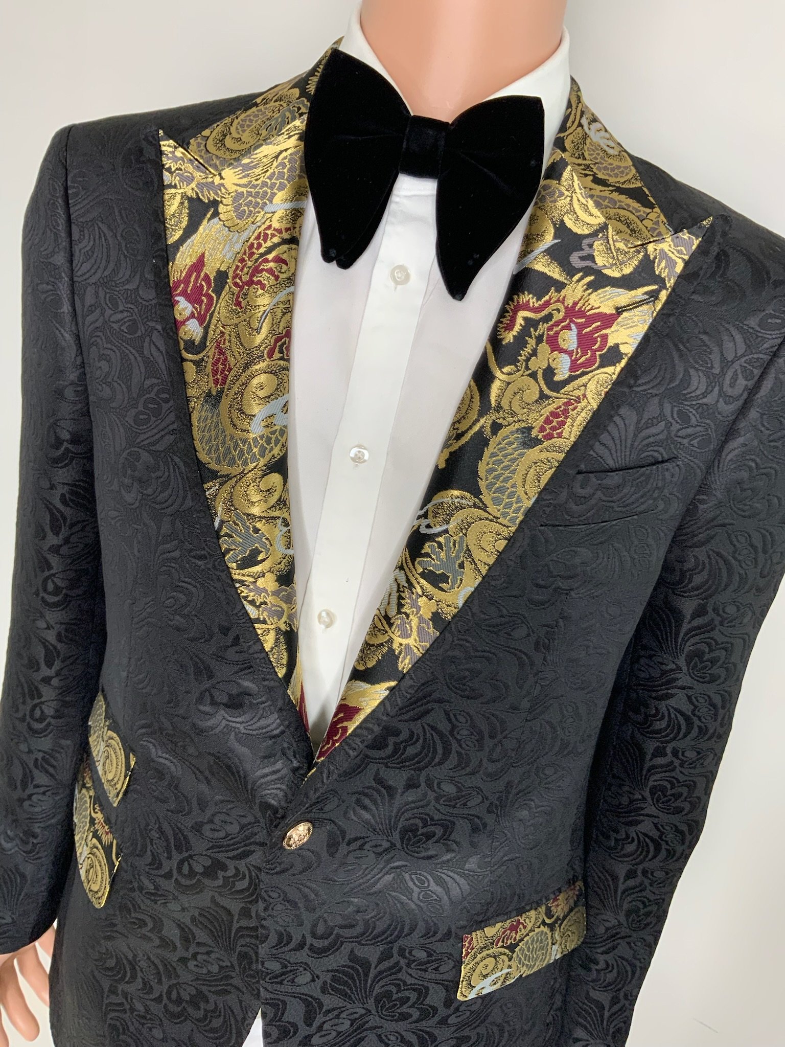 Close-up of a mannequin wearing a black tuxedo with gold embroidered floral designs on the lapels, a white dress shirt with pearl buttons, and a black velvet bow tie.