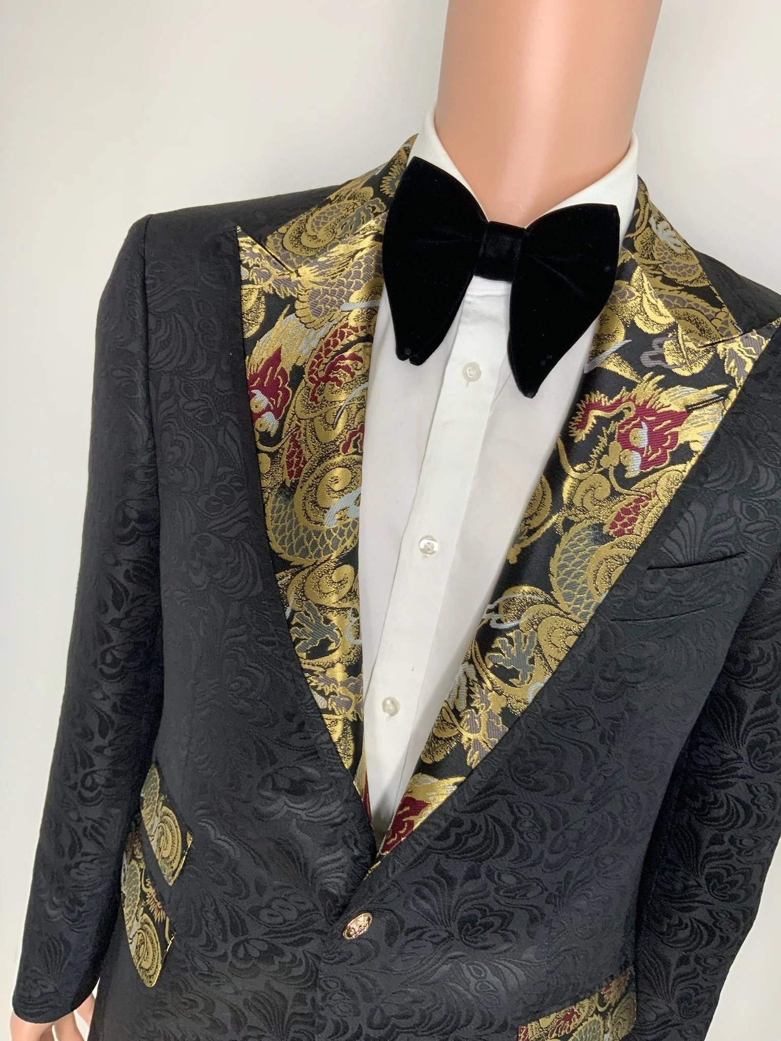 Close-up of a mannequin dressed in a black tuxedo with gold and red floral embroidery on the lapels, a white dress shirt, and a black velvet bow tie.
