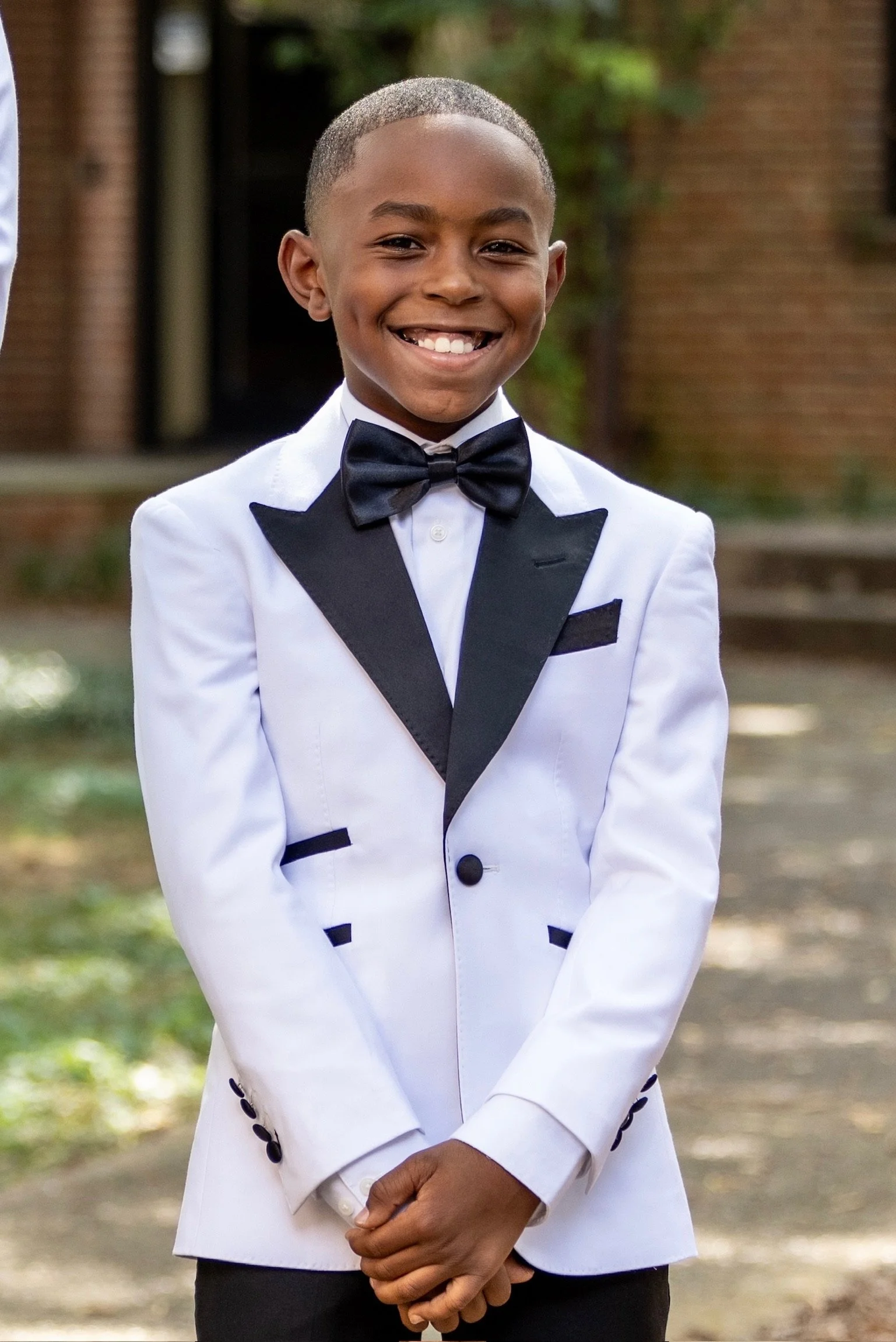 A young boy dressed in a white tuxedo jacket with black lapels and accents, a white shirt, and a black bow tie, smiling outdoors.