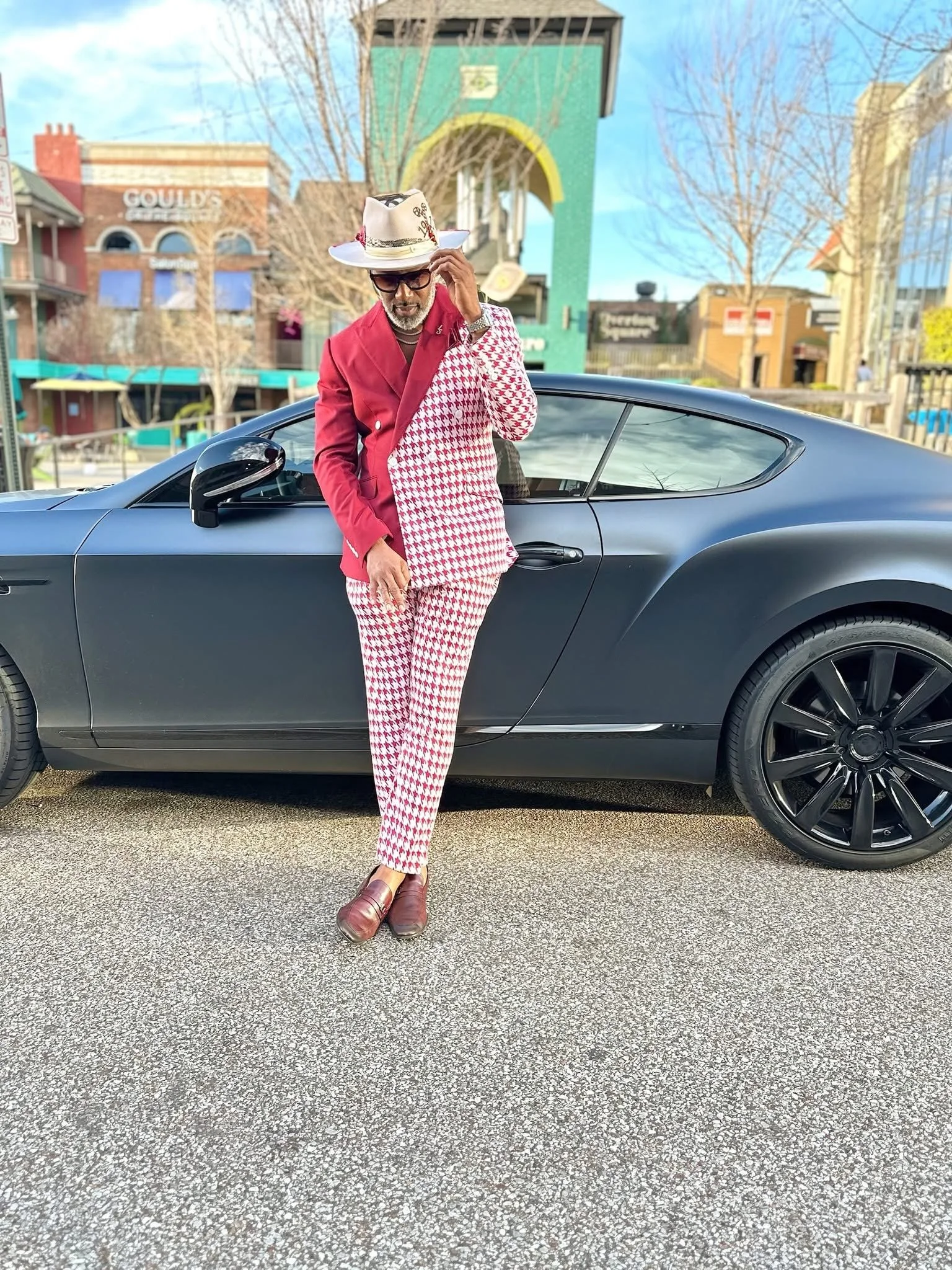 A man dressed in a red and white houndstooth suit, with a red jacket, red and white patterned pants, brown shoes, and a white hat with a red and black band, standing next to a black sports car on a city street. He is adjusting his sunglasses.