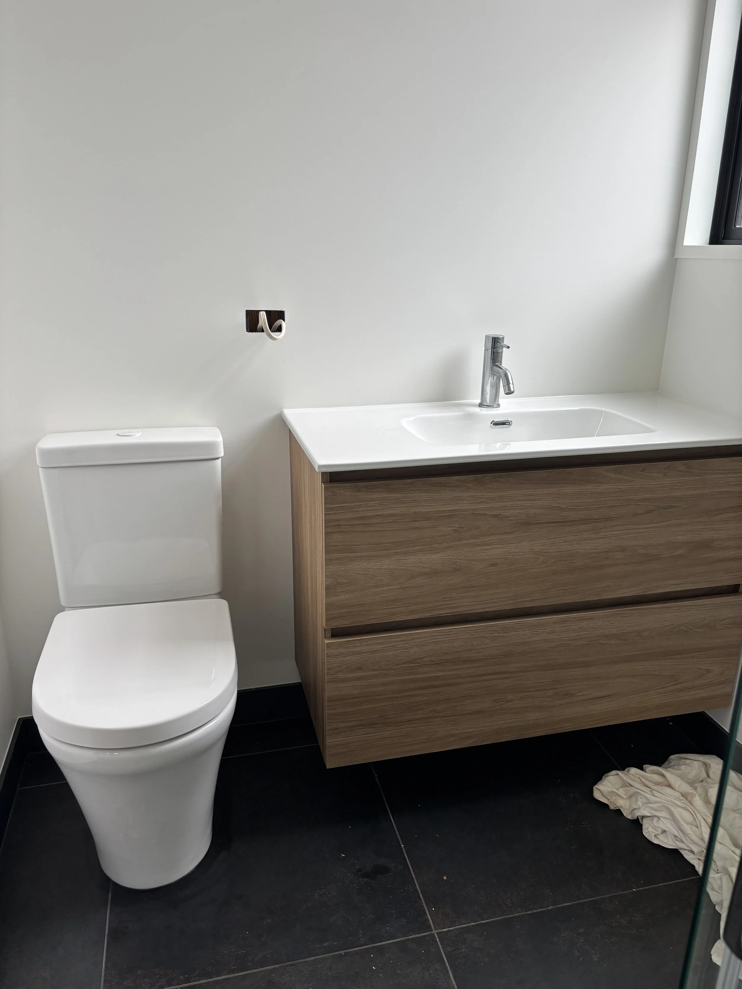 Modern bathroom with white toilet, wooden vanity with white sink, silver faucet, and black tile floor. Cloth on the floor.