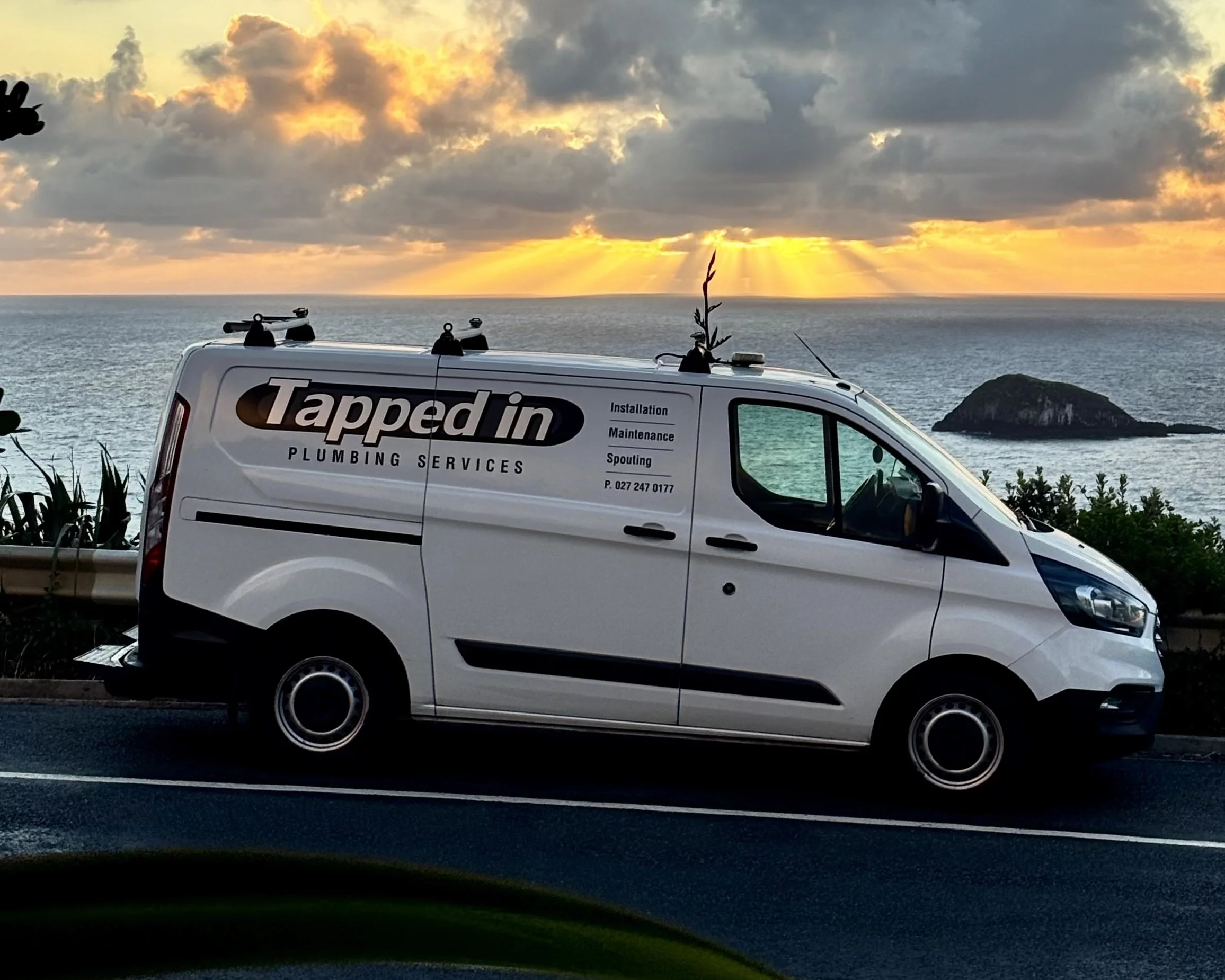 A white plumbing service van parked on a roadside near the ocean during sunset, with a rocky island in the background and a partly cloudy sky with sun rays.