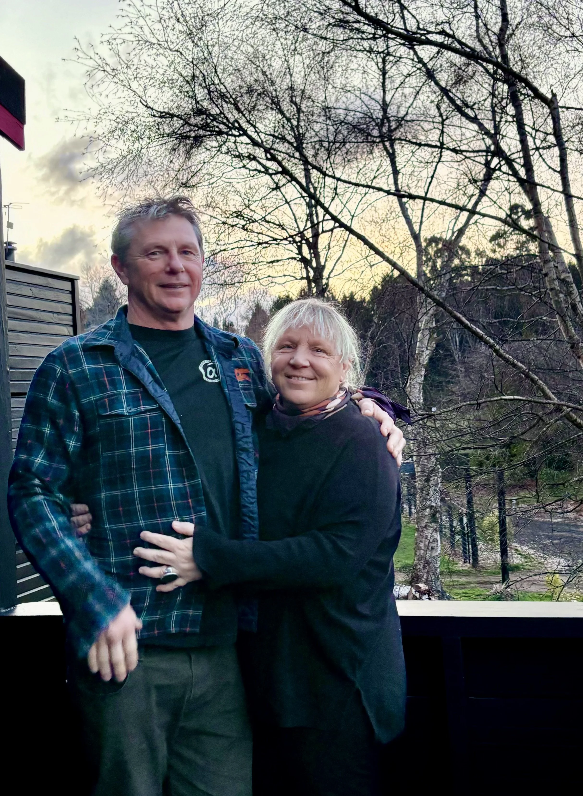 A man and woman standing outdoors on a porch, smiling, with leafless trees and a cloudy sky in the background.