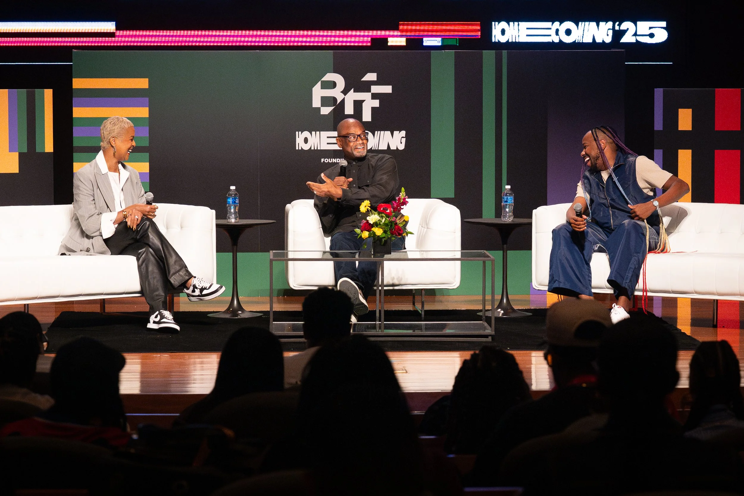 Three people sitting on white couches on a stage during a panel discussion at an event. The person in the middle is holding a microphone and gesturing, while the person on the right is leaning forward with a microphone. A woman on the left is also holding a microphone. There is a table with water bottles and a flower arrangement between the couches. Audience members are visible in the foreground.