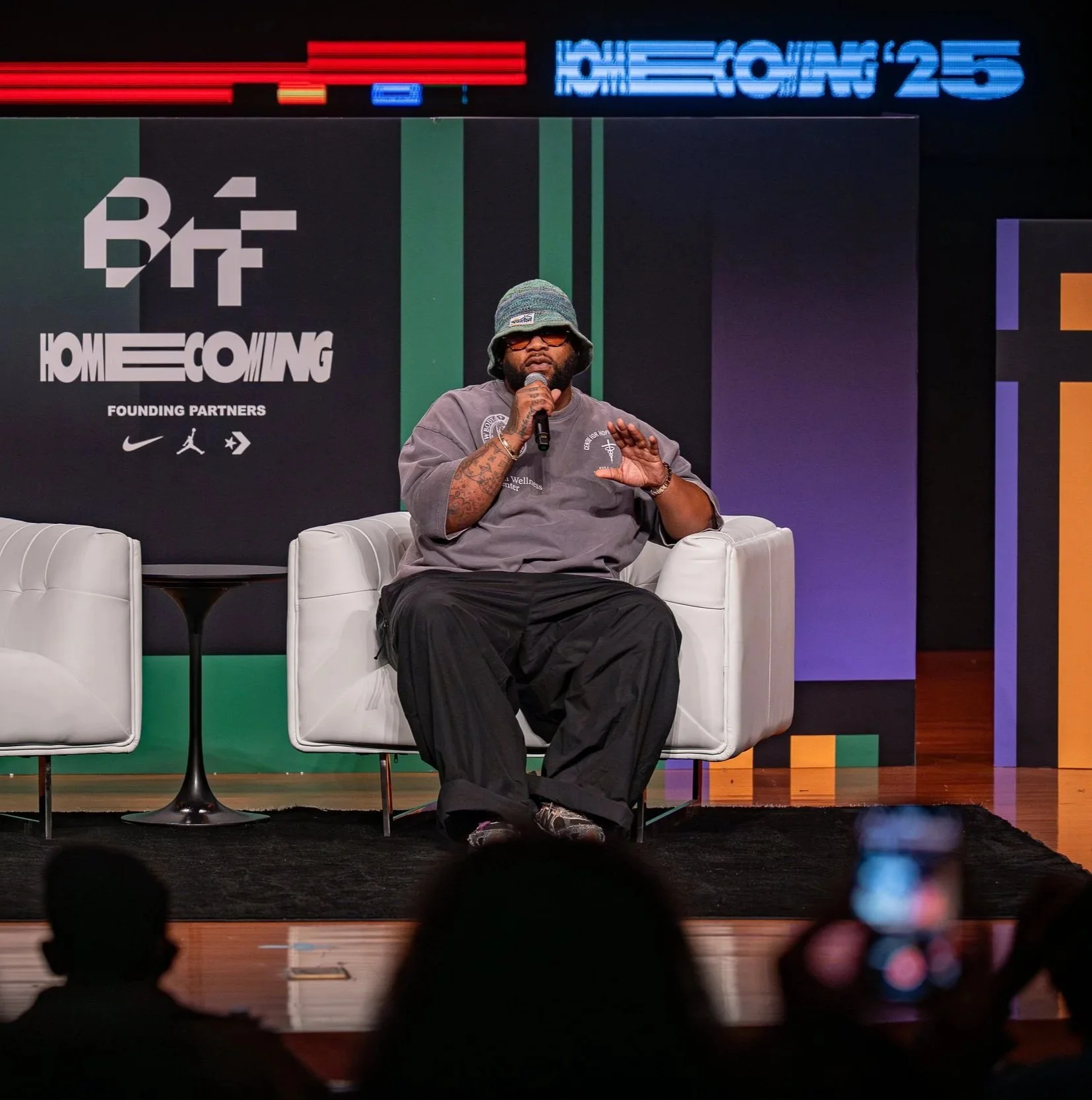 A man with a beard, wearing a bucket hat, sunglasses, a gray hoodie, and black pants, sits on a white armchair on a stage holding a microphone. Behind him is a backdrop with the words 'Homecoming' and logos of Nike and Jordan.