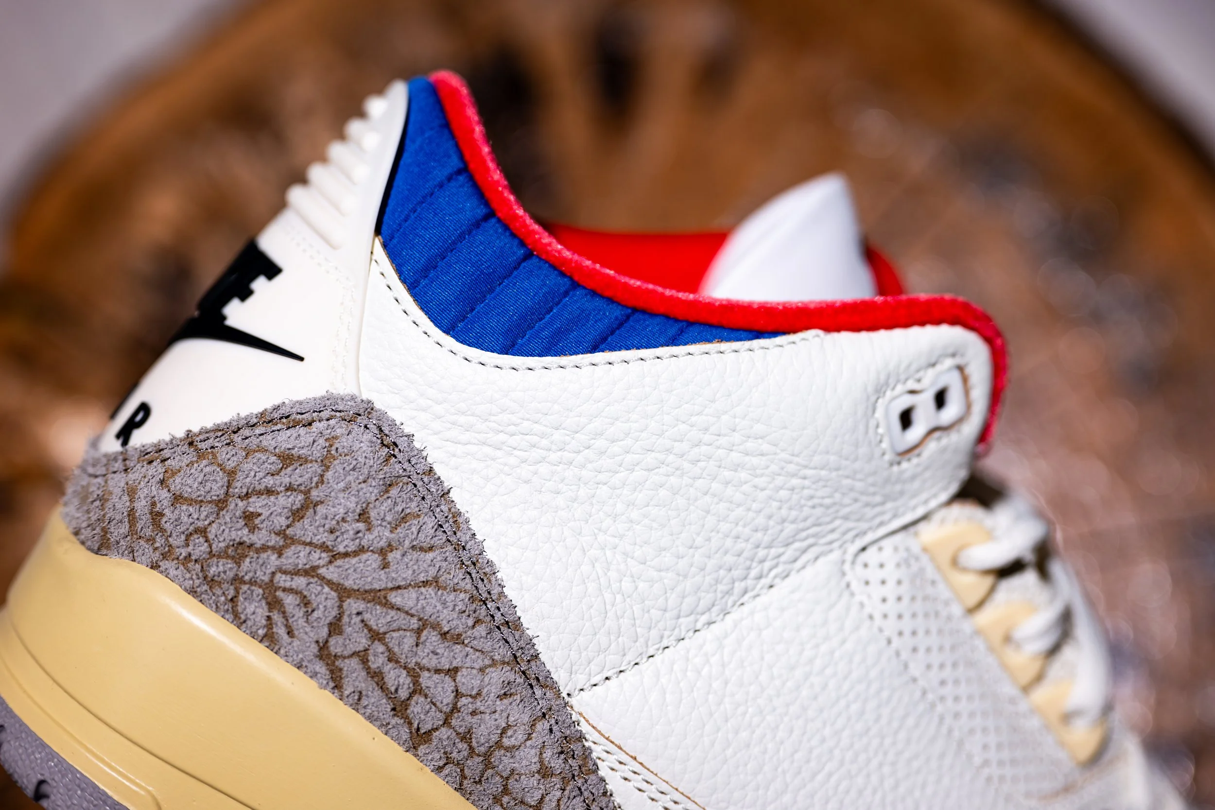 Close-up of a white Nike sneaker with red, blue, and gray accents, showing details of the textured and suede materials, with a focus on the heel and side design.