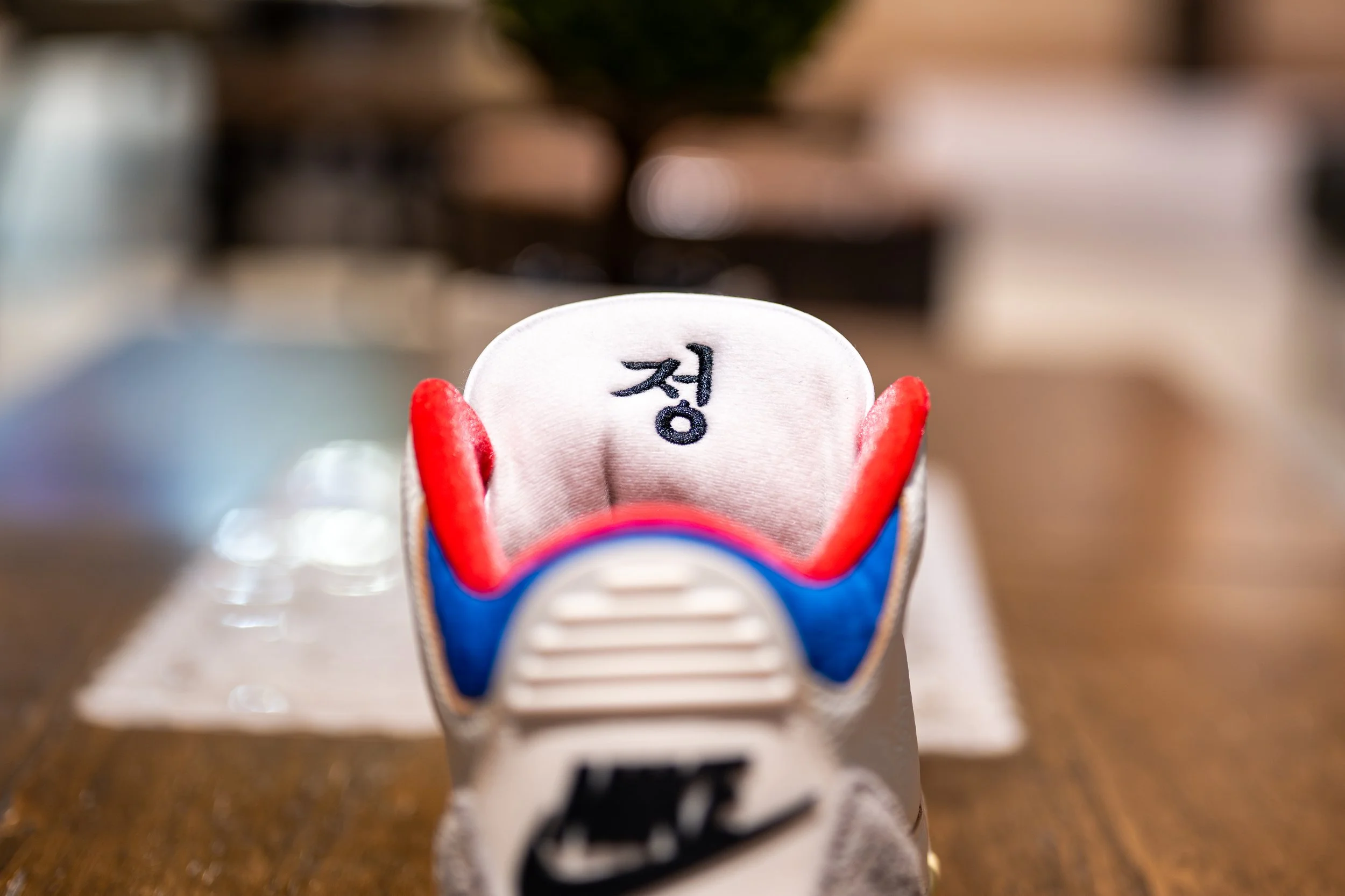 Close-up of a sneaker's tongue with Korean character '정' embroidered on it, featuring red, blue, and white colors, on a wooden table with a blurred background.