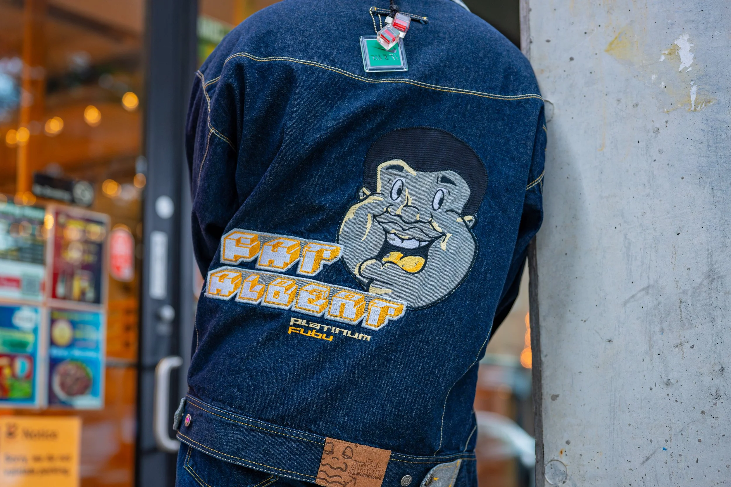 Back of a person wearing a denim jacket with a cartoon face patch and text design, standing near a concrete wall outside a store.
